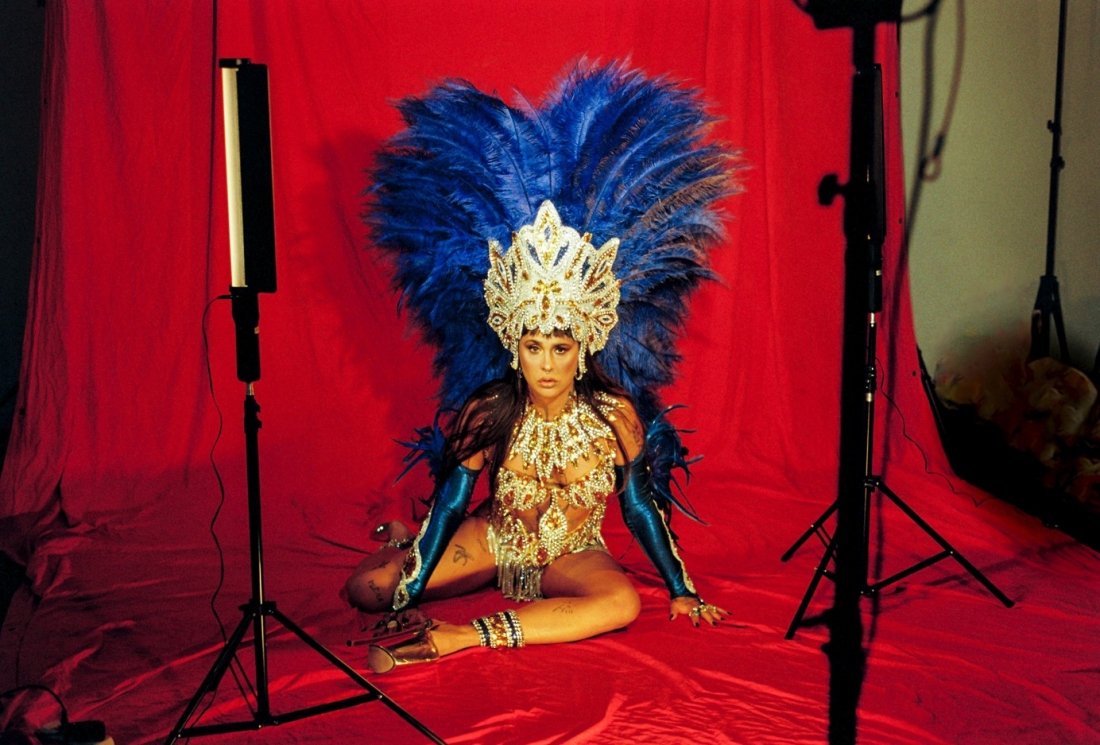 Uma mulher está sentada no chão, diante de um fundo vermelho, usando um traje típico do Carnaval do Rio com penas azuis e acessórios brilhantes. A cena é enquadrada por equipamentos de iluminação visíveis, sugerindo um ambiente de estúdio fotográfico.