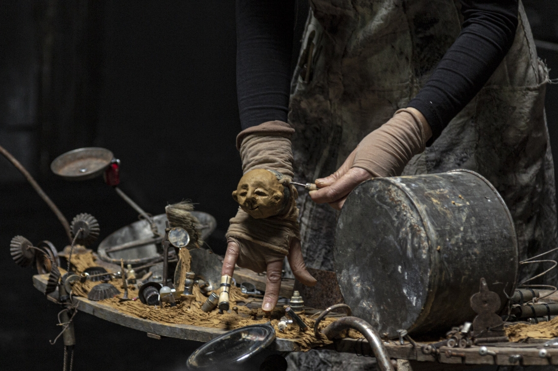 Colour stage photo. In a close-up, Sandra Neves makes her hand a puppet with a clay head. The hand made the puppet walks in a landscape of metal spoils.