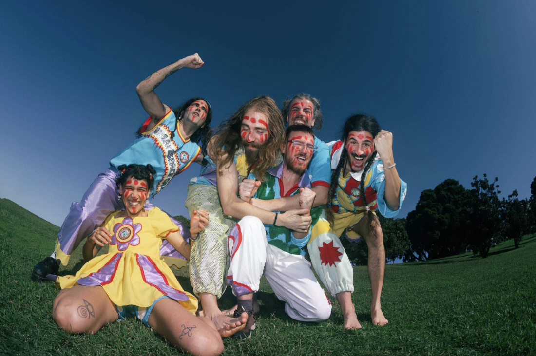A group of six people pose, smiling, outdoors on a lawn, wearing colourful costumes and red face paint.