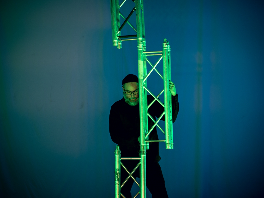 A man is holding a vertical metal structure, illuminated by green light, against a blue background.
