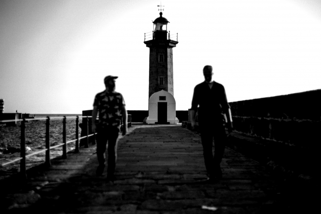 Black and white photo. On a pier, with their backs to a lighthouse, Afonso Dorido and Francisco Silva walk towards the camera.