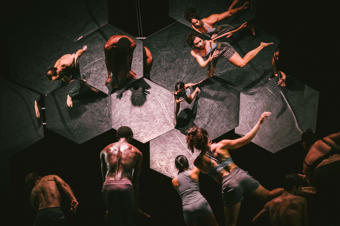 Colour stage photo. Performers with their backs to the audience and facing a mosaic of hexagonal mirrors, which reflects their bodies.