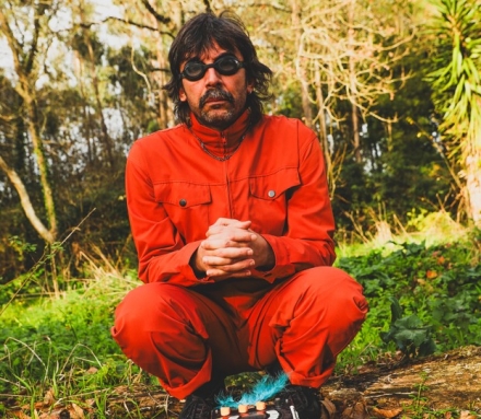 Retrato a cores de Franklin Soares Monteiro. Está agachado numa floresta, com um macacão laranja, com equipamento de música no chão à frente.