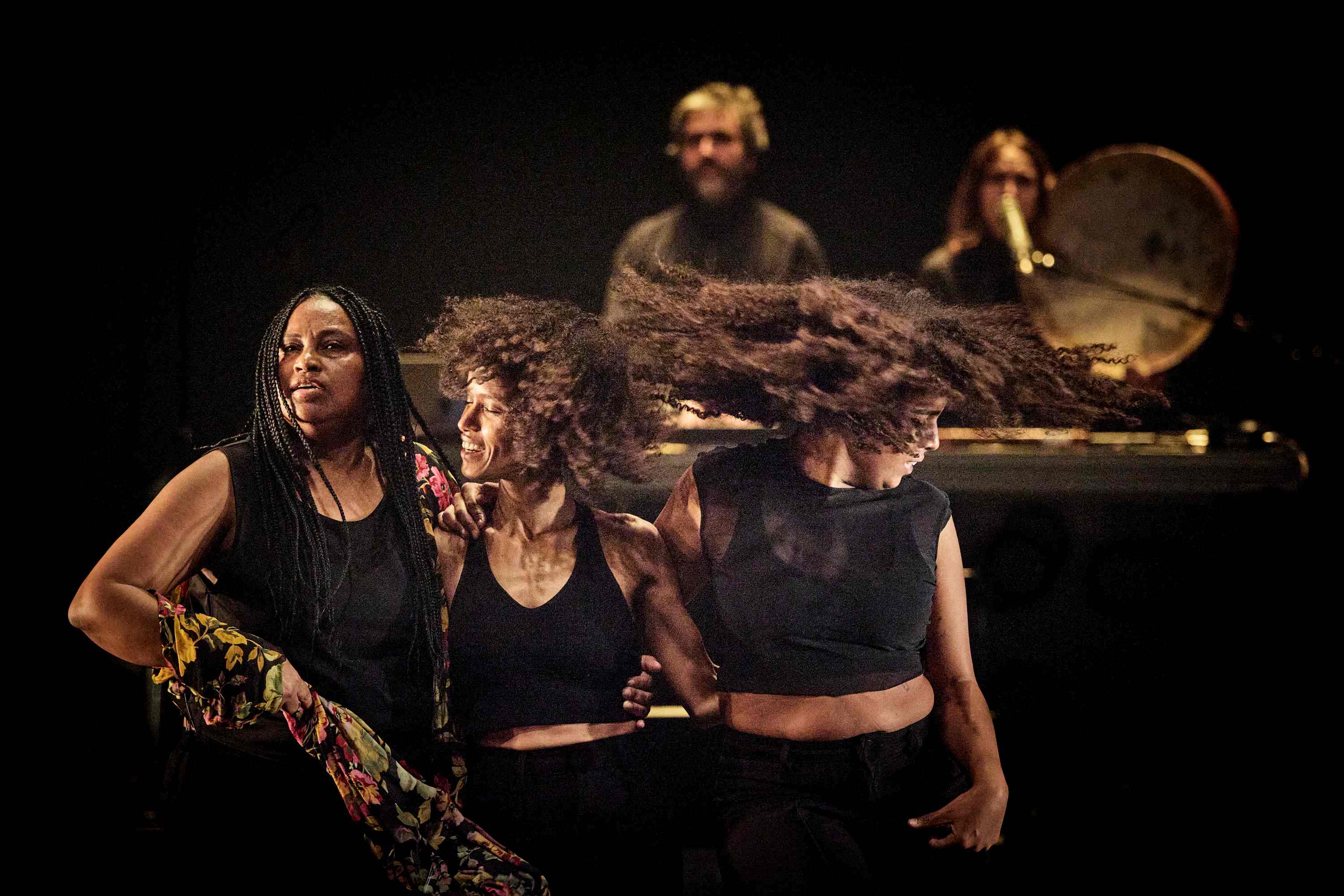 Three performers, standing side by side on stage, turn their heads, with musicians in the background.