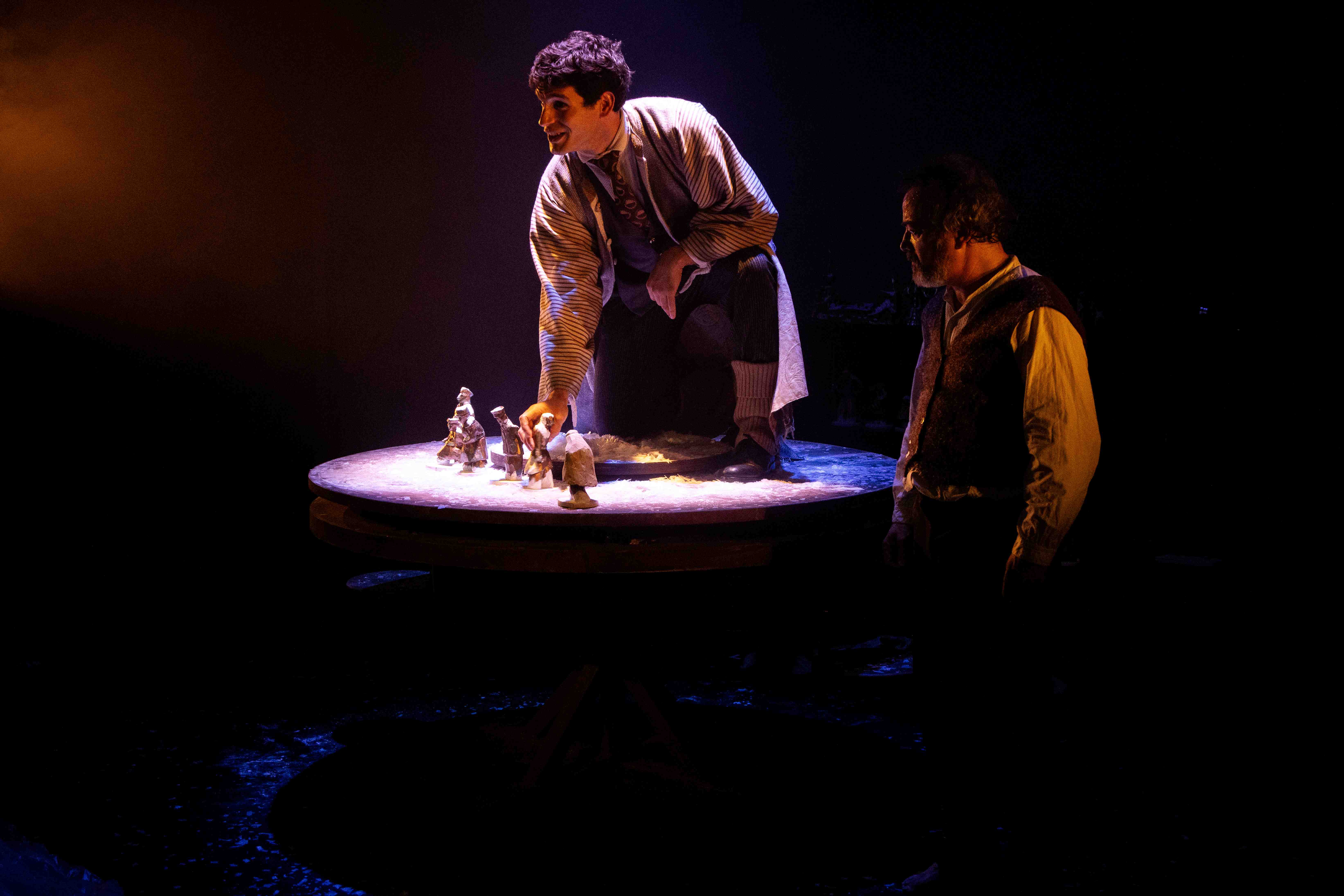 Two actors manipulate small figures on an illuminated table.