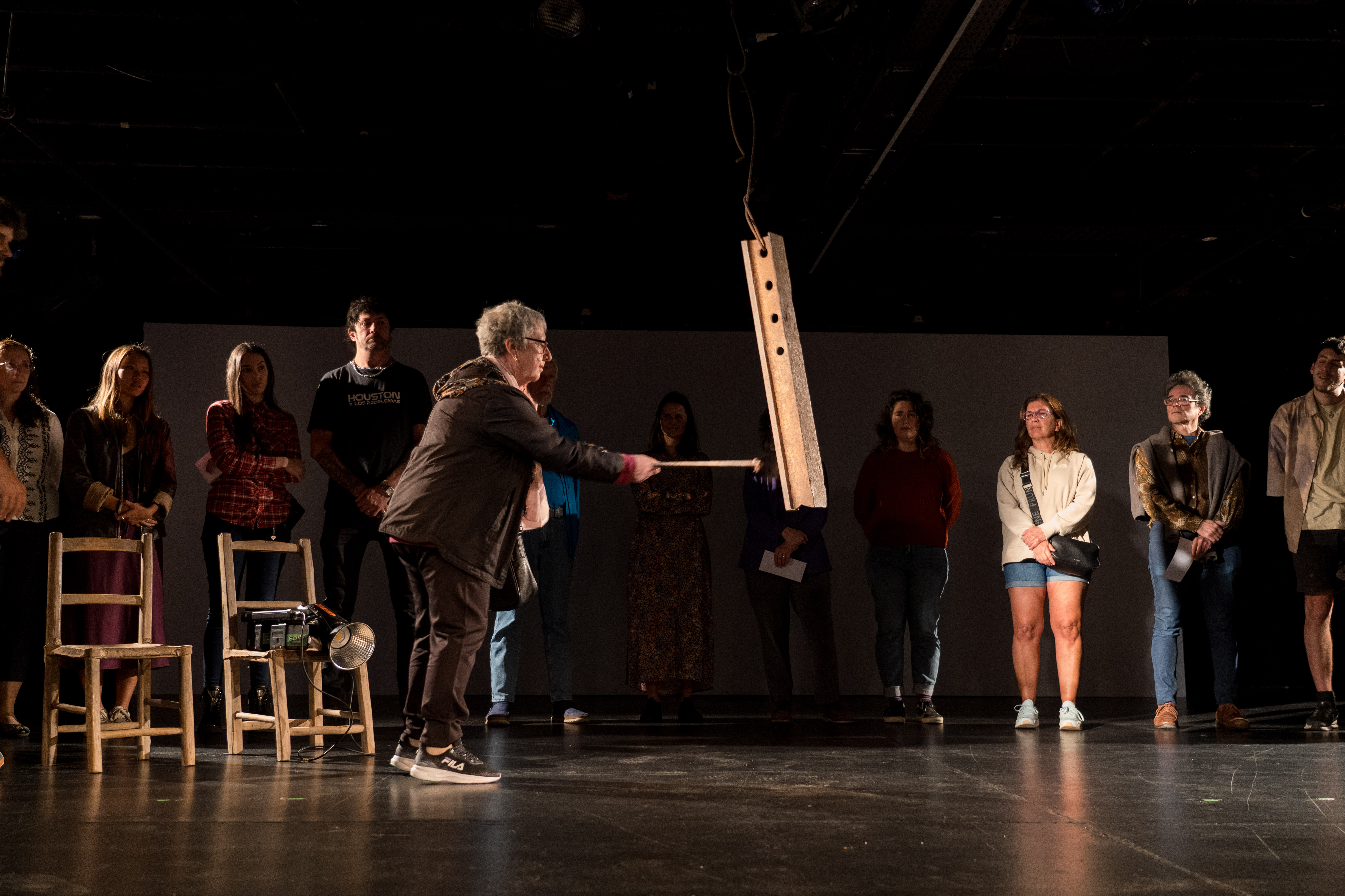 Num palco, rodeada por várias pessoas, uma mulher bate com uma vareta numa peça de metal suspensa.