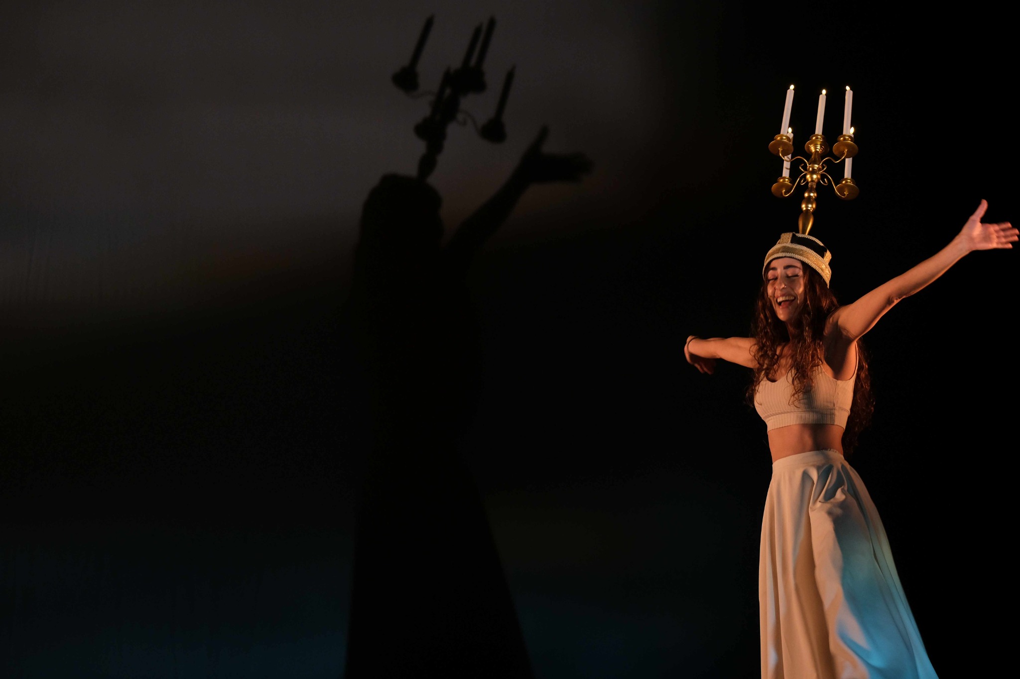 A performer wearing a candelabrum with lit candles on her head casts a large shadow across the stage.