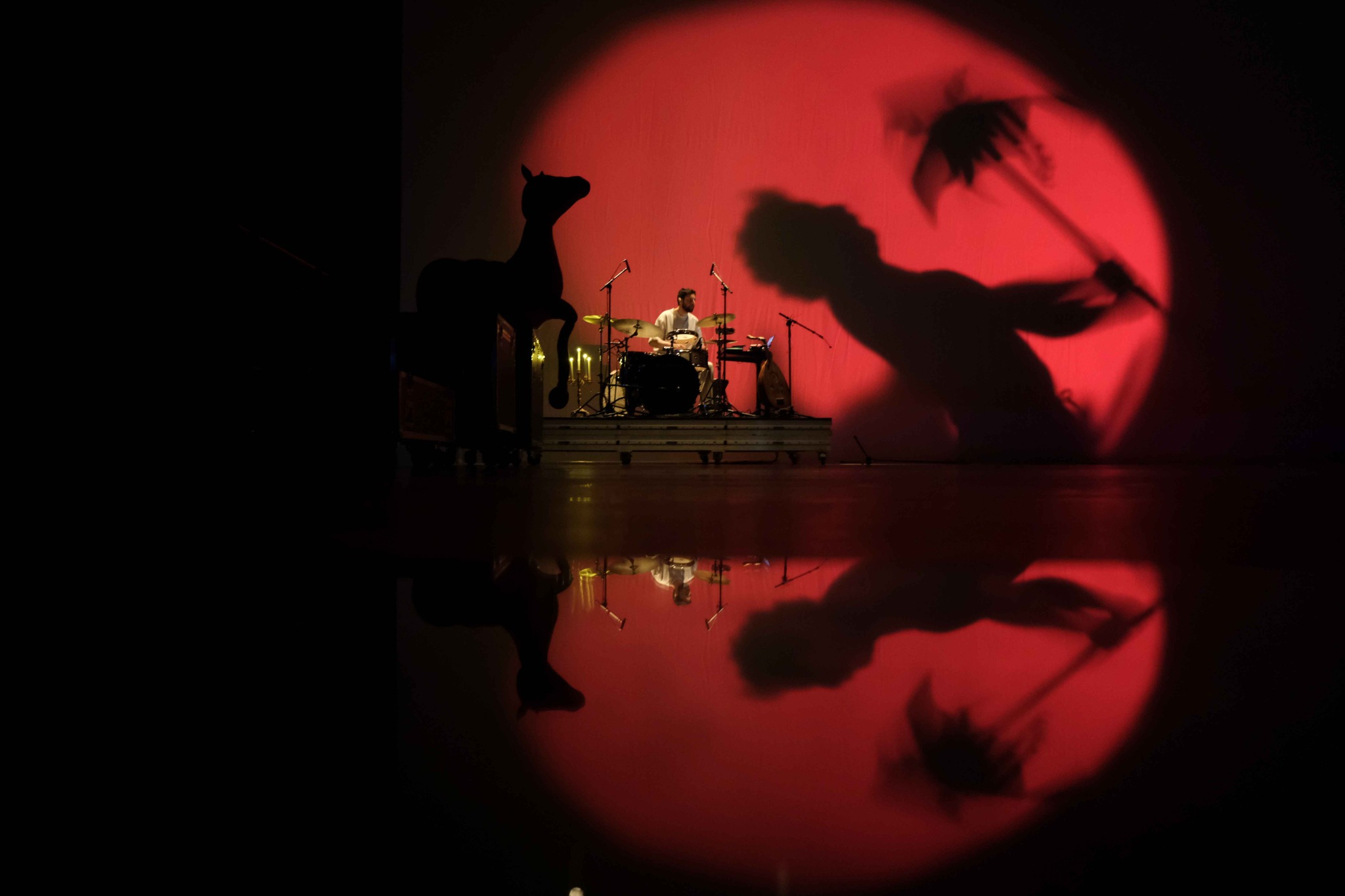 An enlarged shadow of a figure holding an object is cast onto a red circle, whilst a drummer plays in the background.