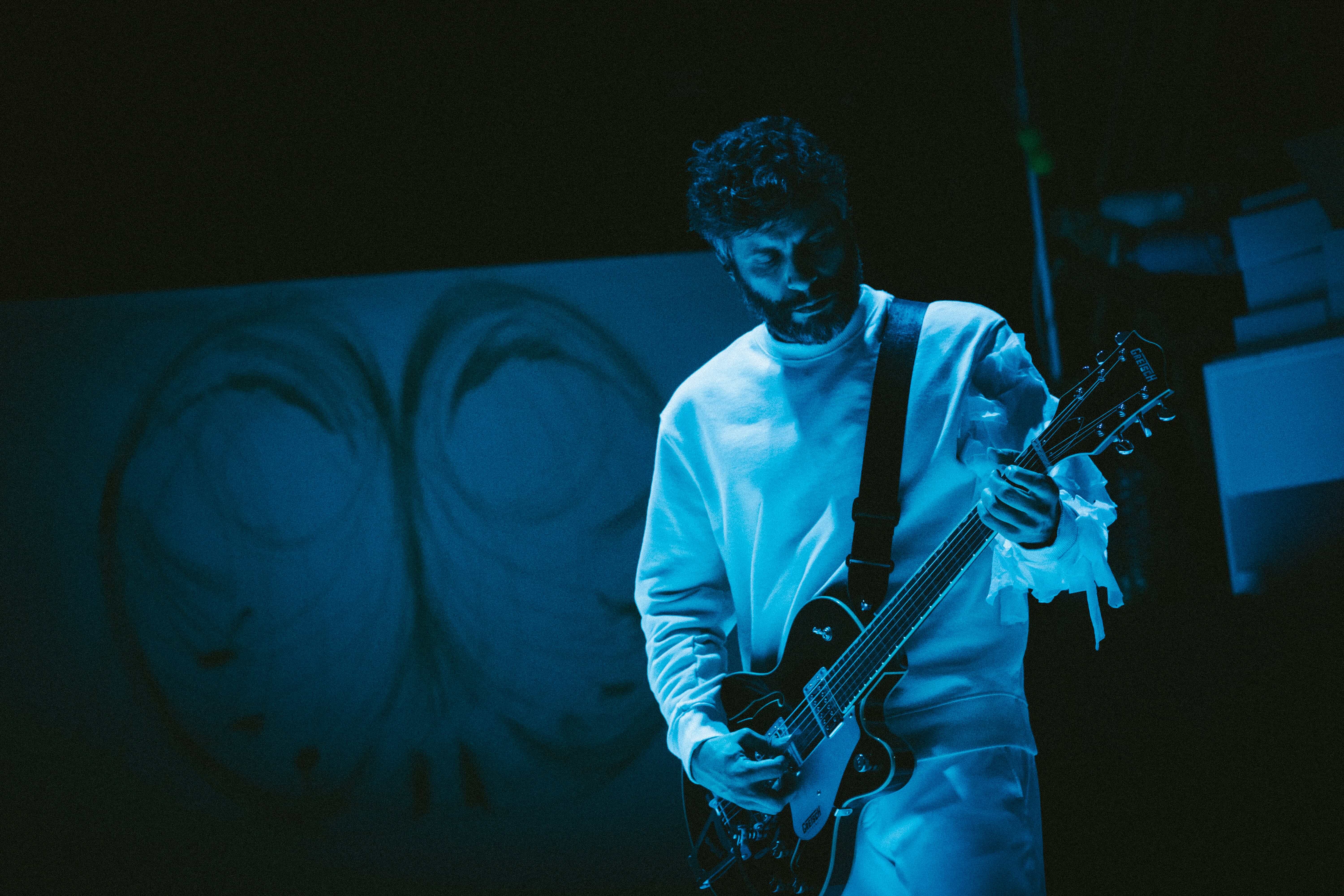 Fotografia de cena a cores com filtro azul. Músico, vestido de branco, toca guitarra elétrica.