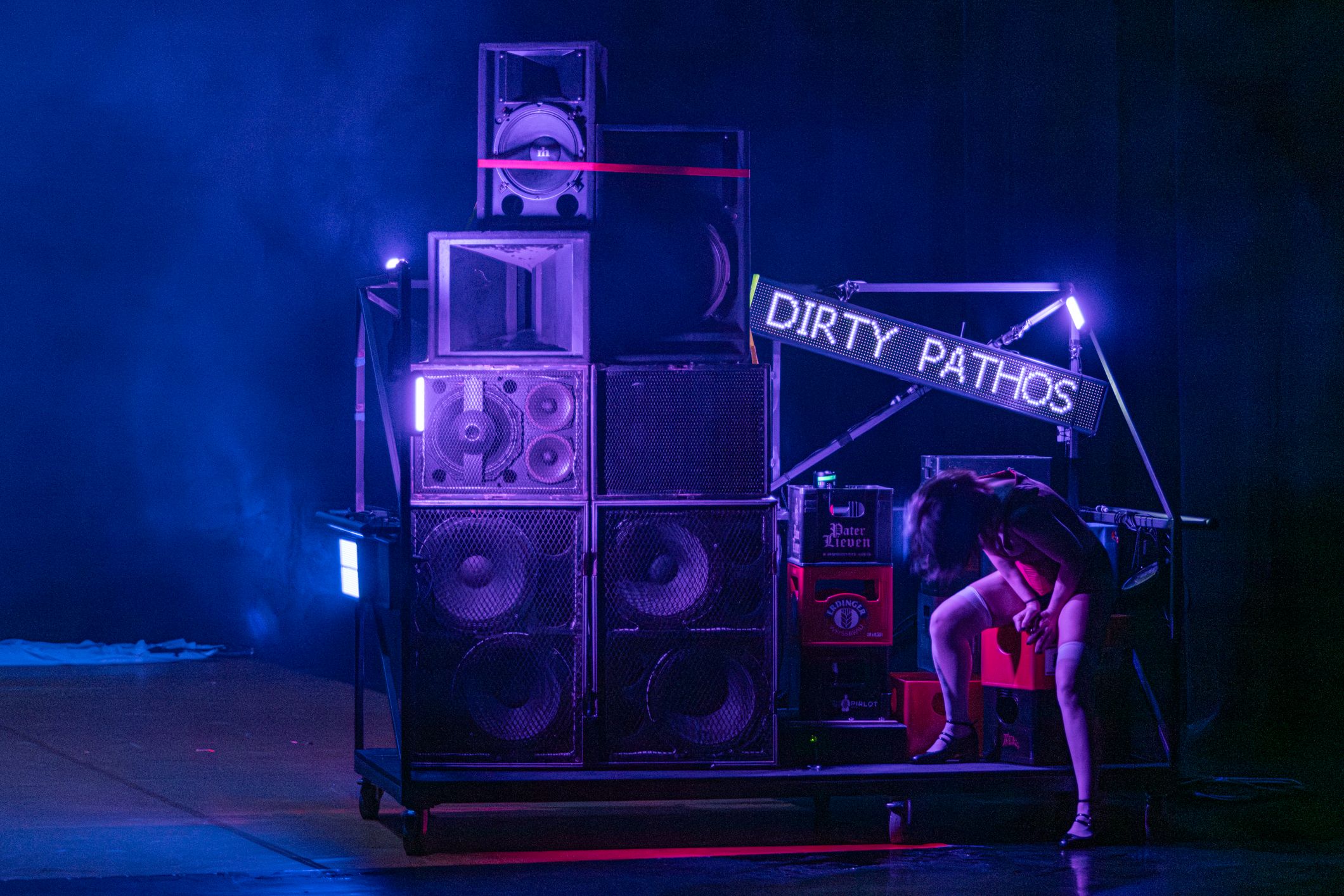 Next to a structure featuring loudspeakers and a neon sign reading ‘DIRTY PATHOS’ on stage, a performer bends over, bathed in purple light.