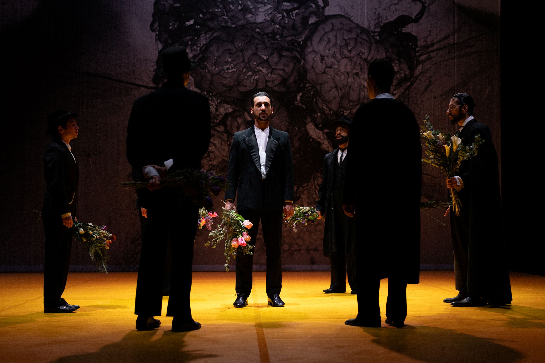 Six performers dressed in black form a circle on stage, holding bouquets of flowers, with a man in the centre standing in front of a large backdrop.