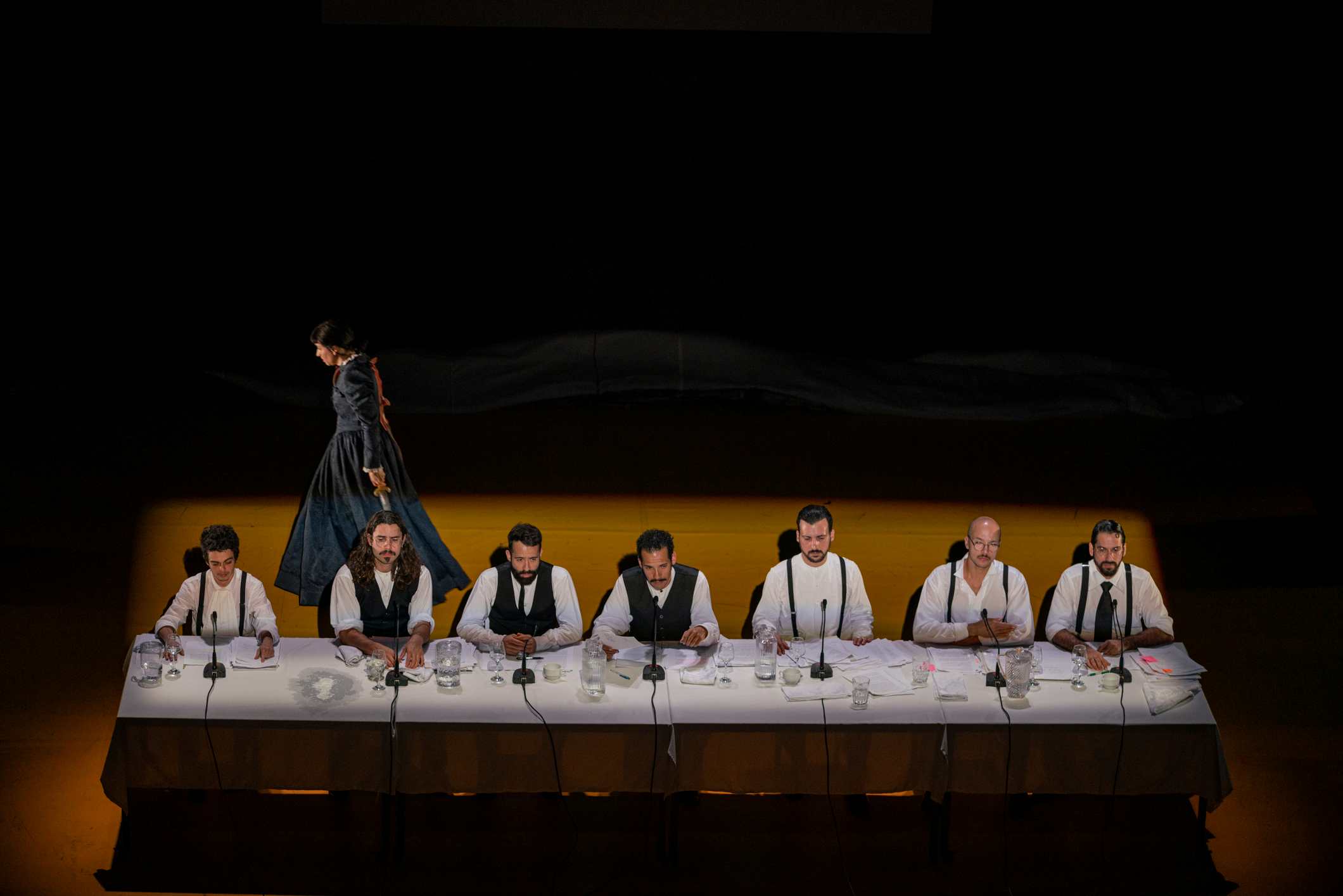 Seven men are seated at a long table with microphones, whilst a woman in a dark dress walks towards the back of the stage.