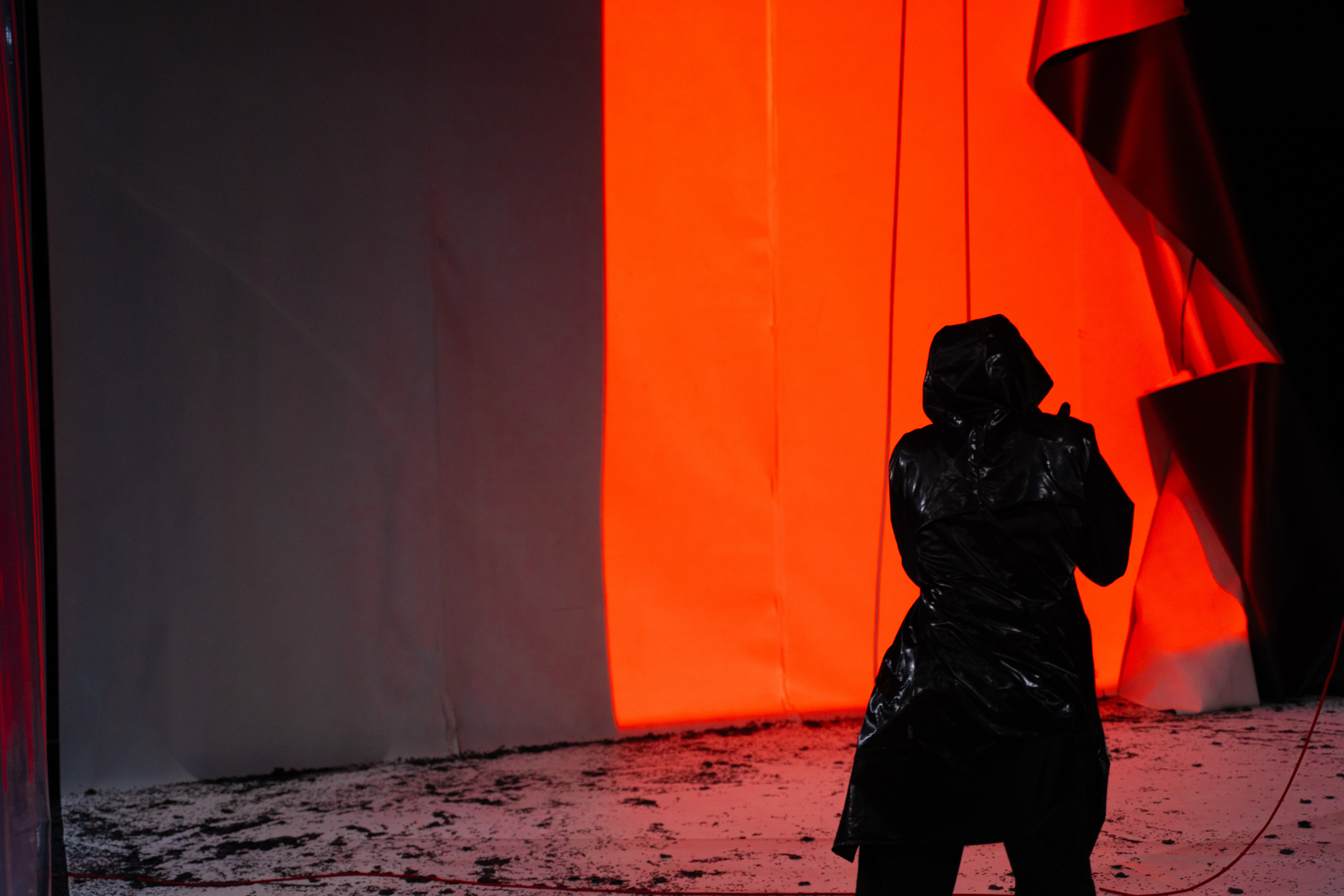Fotografia de cena a cores. Uma ntérprete, de costas, num palco sujo. Ao fundo, uma luz intensa laranja projetada numa tela. 