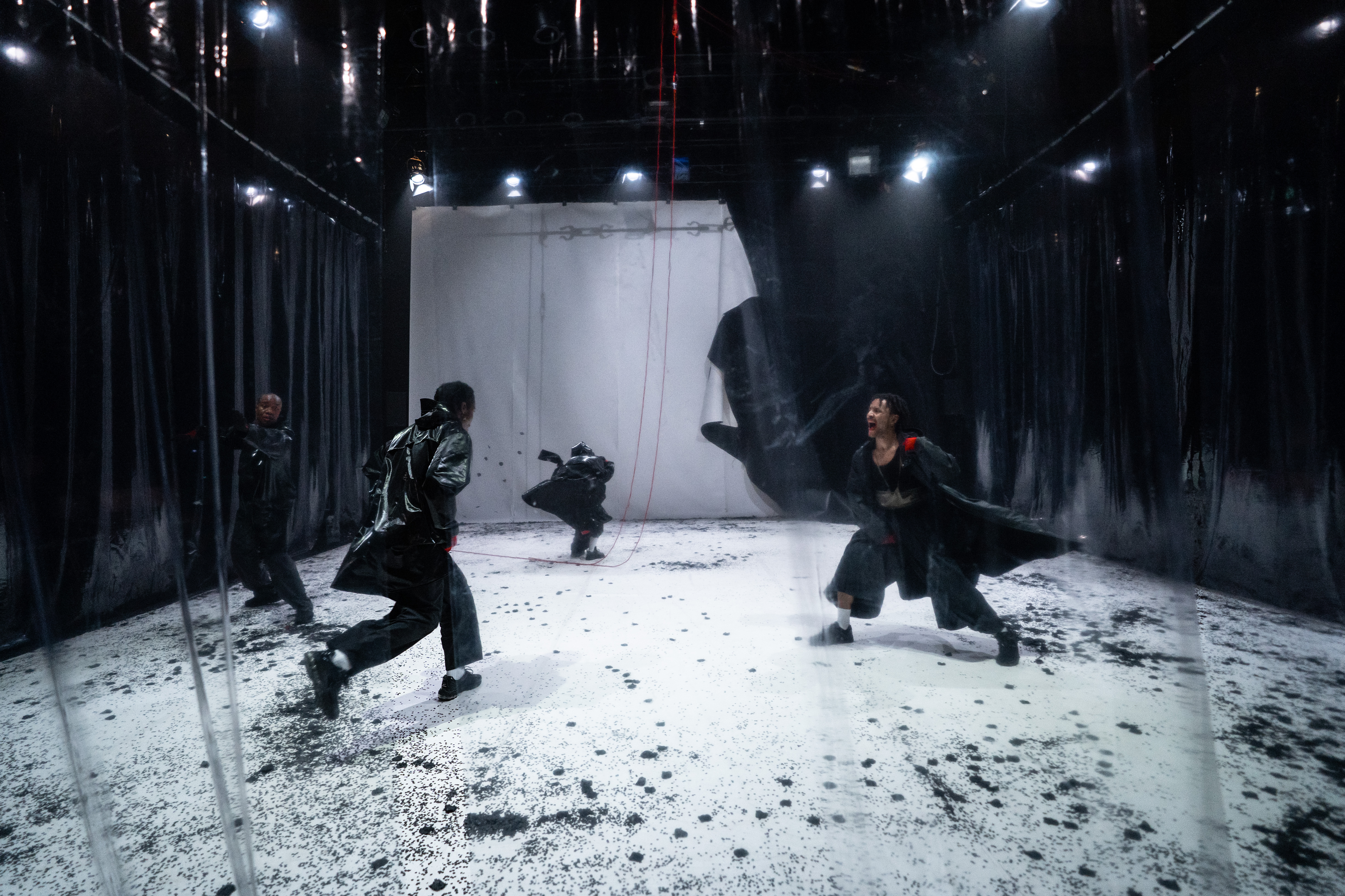 Fotografia de cena a cores. Num palco envolto em telas de plástico transparente e preto, quatro intérpretes, todos de preto, movimentam-se em diferentes sentidos.