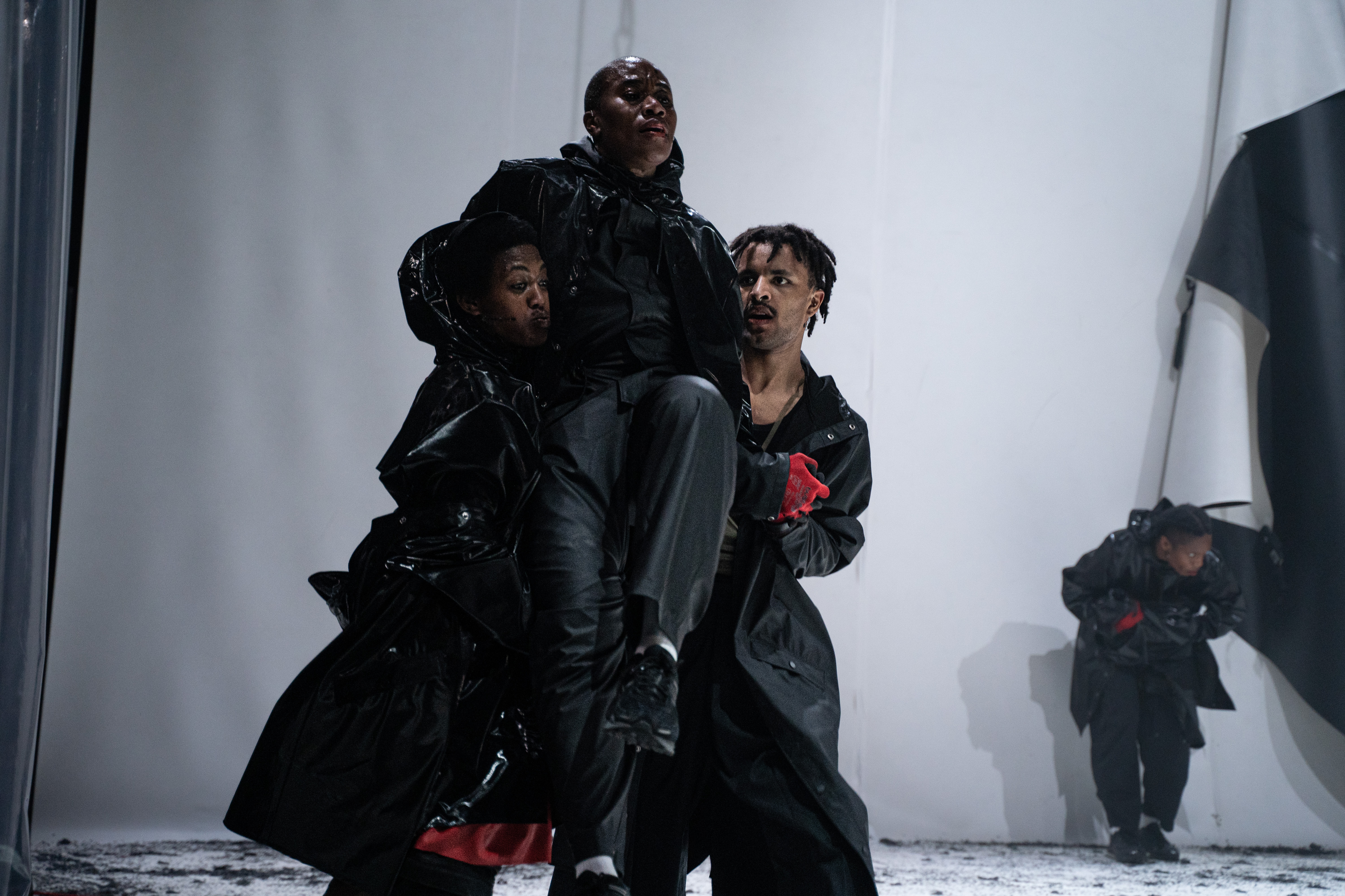 Fotografia de cena a cores. Dois intérpretes elevam um terceiro intérprete, estão vestidos de preto e luvas vermelhas.