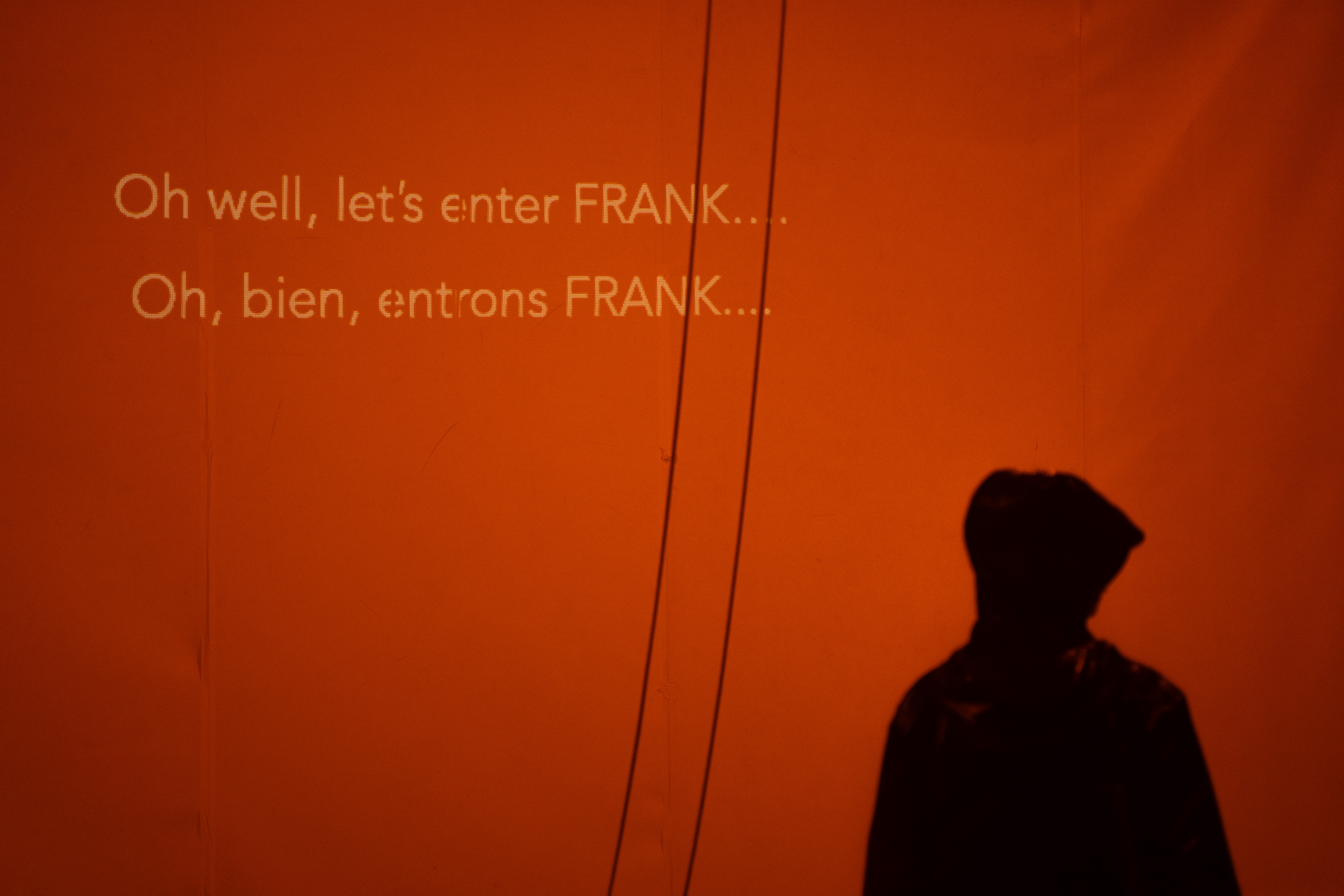 Fotografia de cena a cores. A silhueta desfocada de um intérprete encapuçado, atrás deste, uma tela laranja com legendas projetadas em inglês e francês.