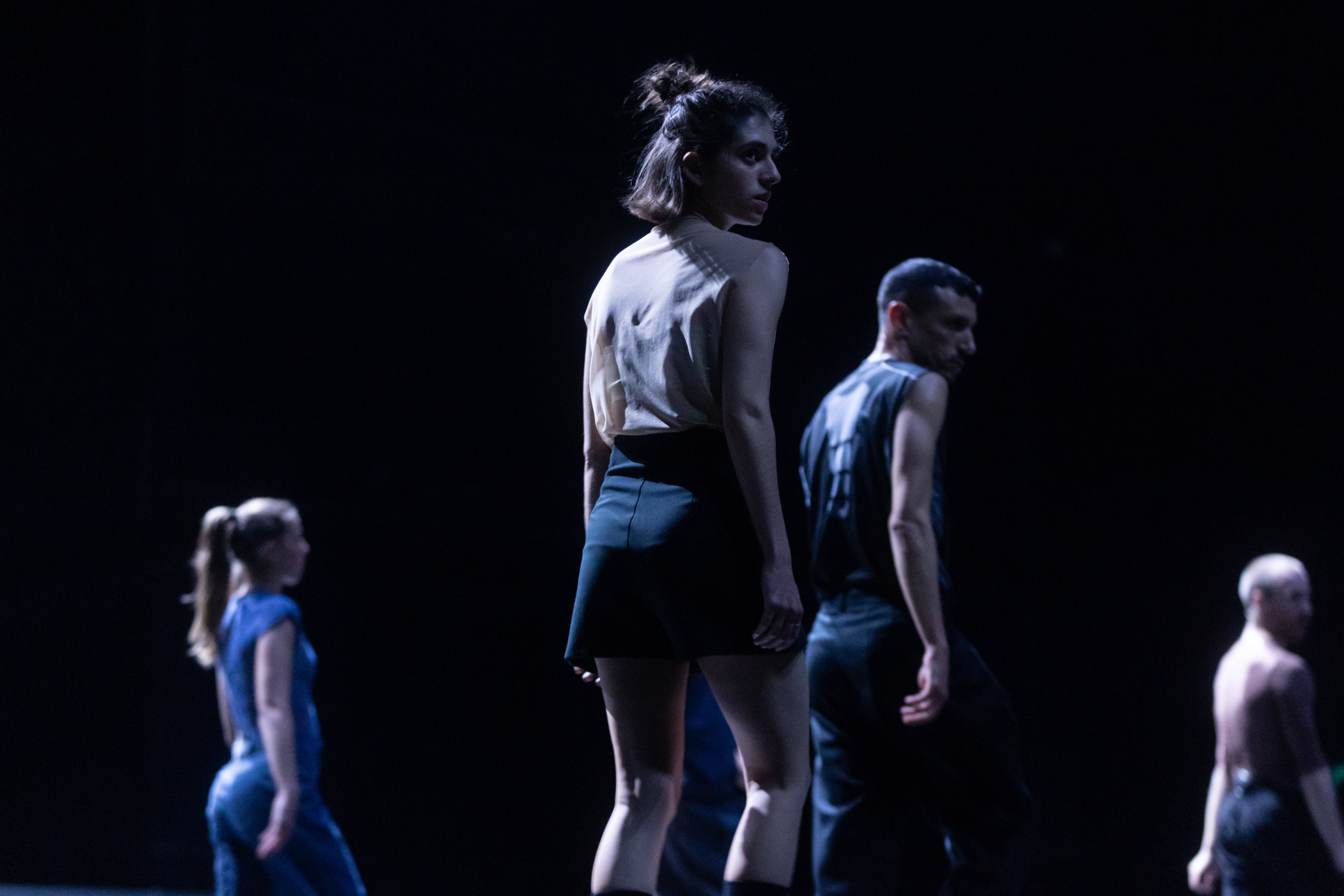 olor stage photograph of a performer in profile, turning her face over her shoulder toward the audience, while other performers in the background mirror the same gesture.