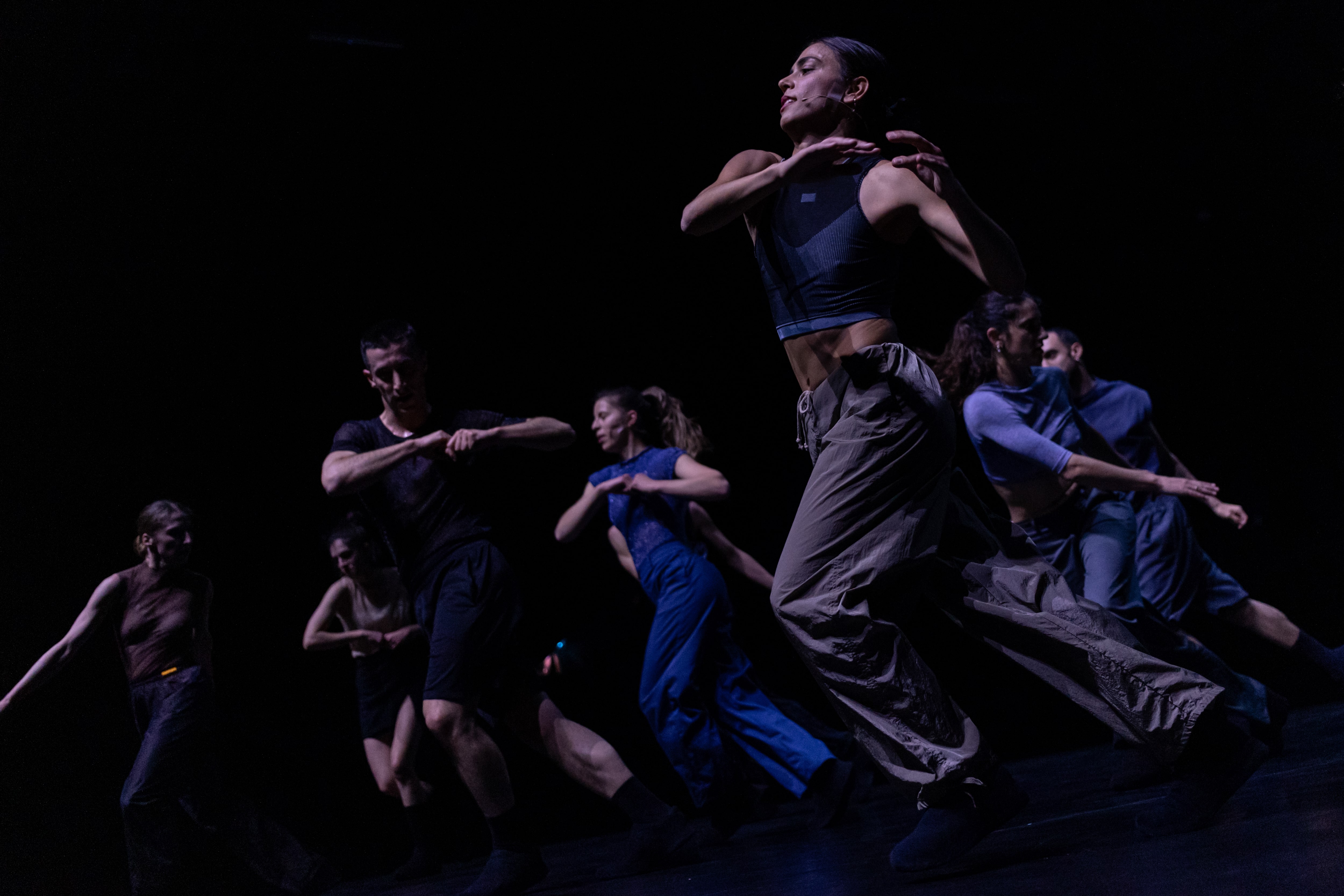 Color stage photograph of a group of performers moving together to the other side of the stage, twisting their torsos in the opposite direction of their hips, with their arms sometimes bent following the torso’s movement.