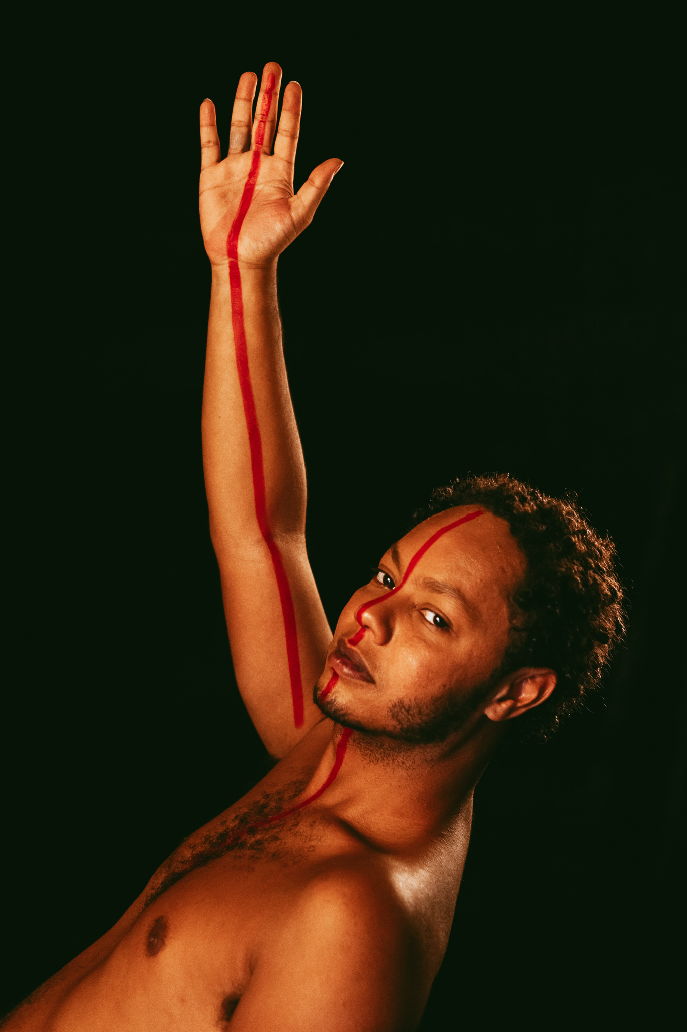 A portrait of Clayton, bare-chested, lit against a black background, with his left arm raised and his palm facing forwards. A painted red line runs across his palm, arm, face and chest.