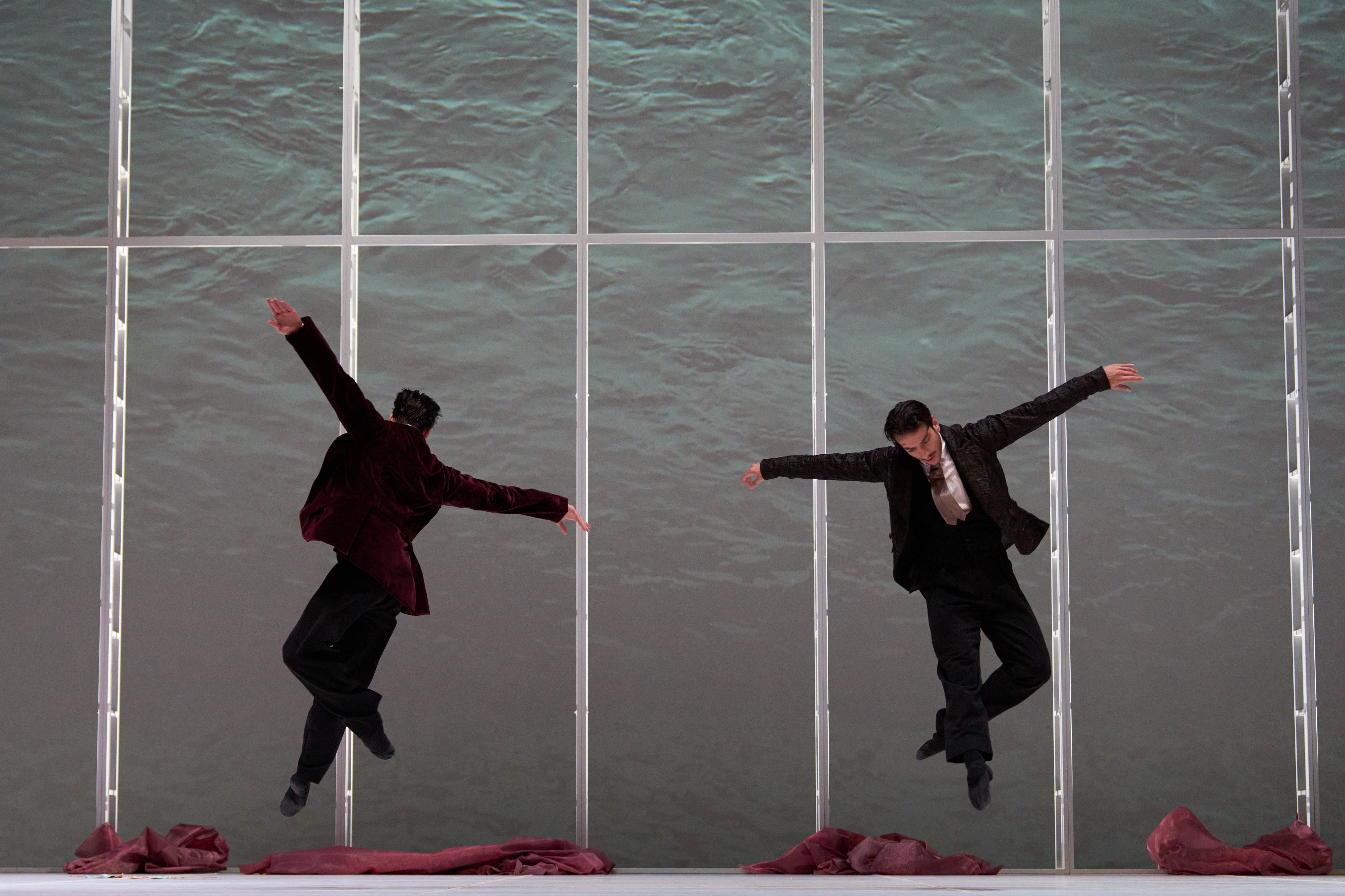 Color stage photograph. Two performers in dark jackets leap simultaneously with open arms in front of a backdrop resembling moving water; reddish fabrics rest on the floor.