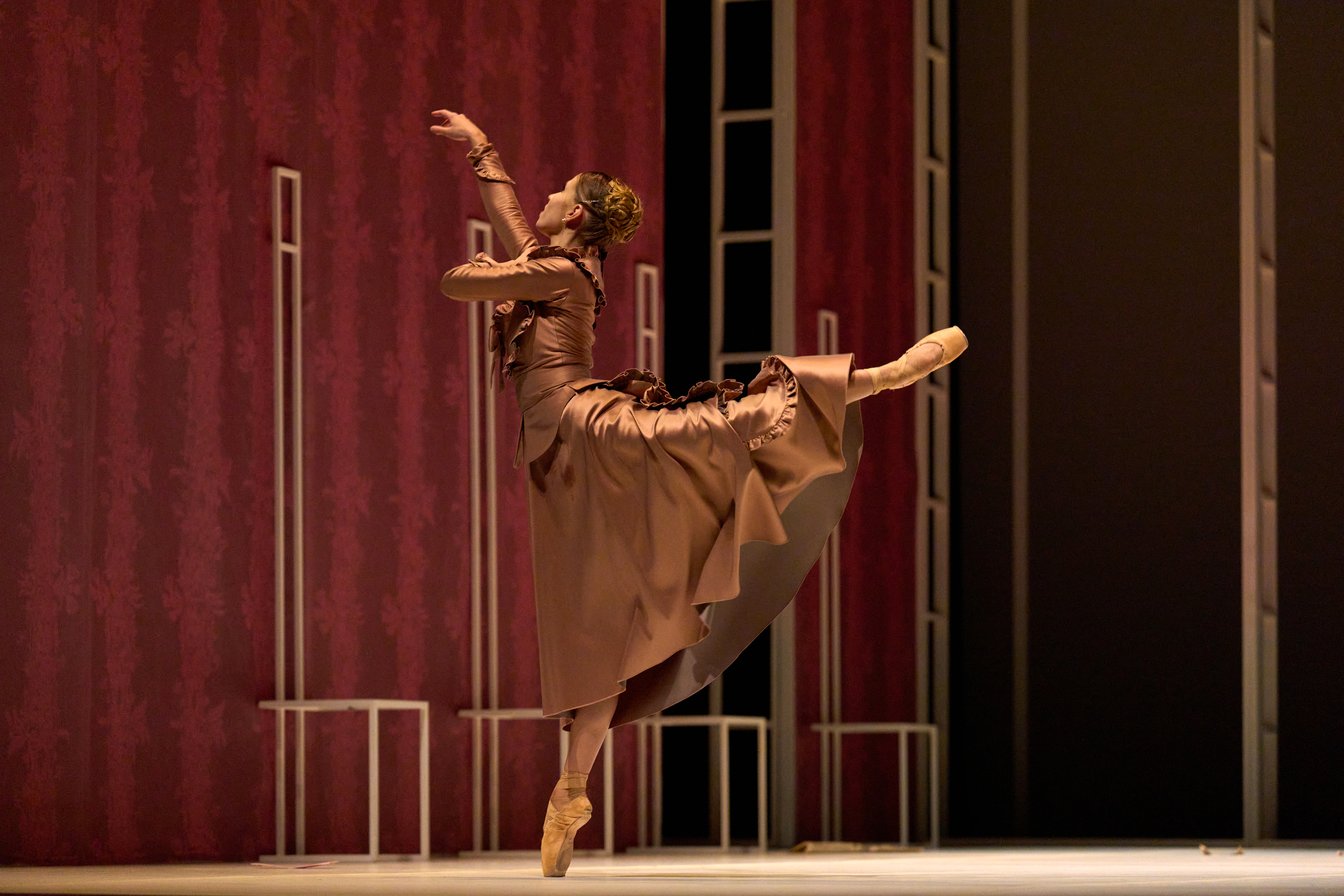 Color stage photograph. A ballerina in a long dress and on pointe lifts one leg while extending her arms, set against a textured red backdrop.