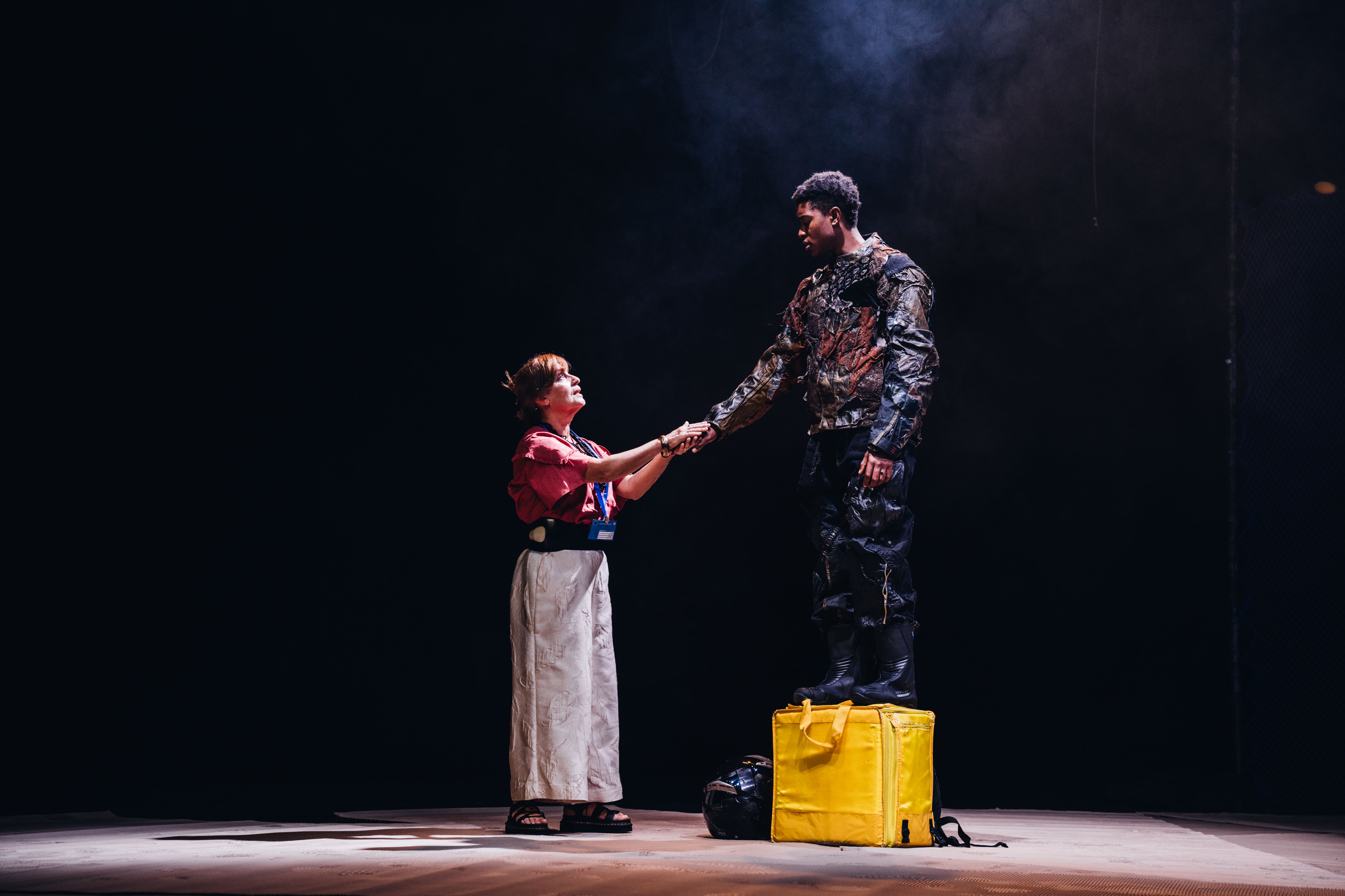 Uma mulher branca com uma credencial azul de uma instituição europeia segura a mão a um homem negro, em cima de uma mochila térmica de entregas de alimentos amarela. Ele e ela olham-se nos olhos.