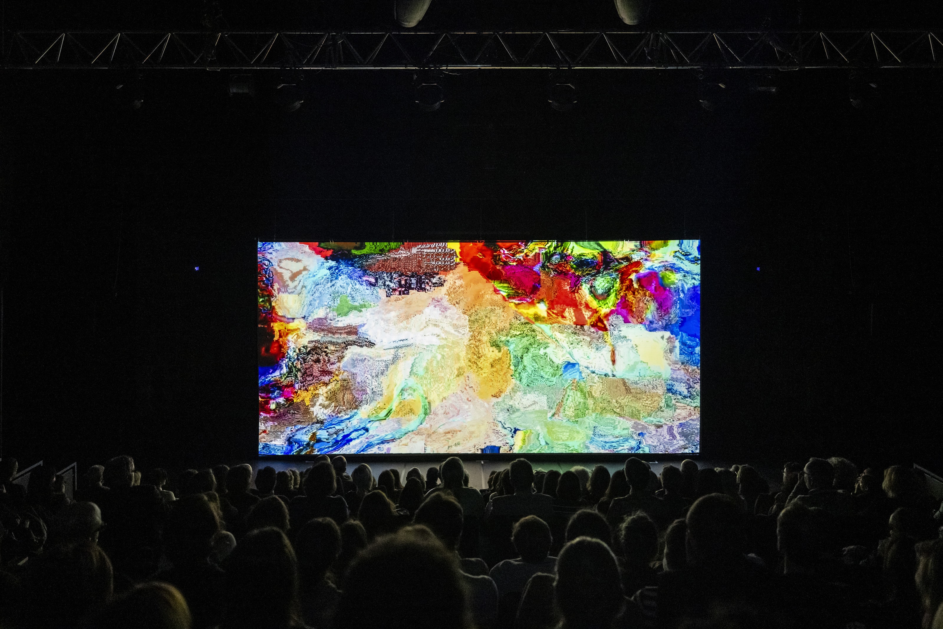 Fotografia de cena a cores. Numa sala com a plateia cheia de pessoas, uma pintura abstrata e colorida é projetada numa tela. 