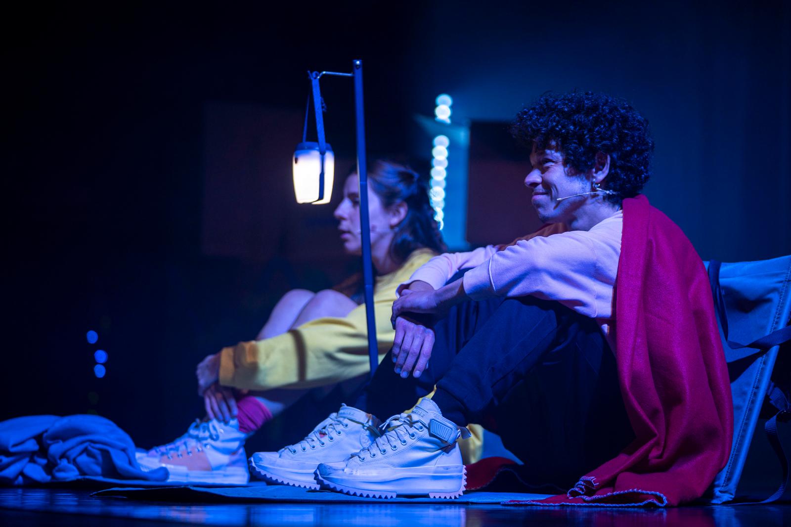 Duas pessoas estão sentadas no chão de um palco, sob luz azul, com expressão tranquila. Uma delas usa um microfone de cena. Entre elas há um candeeiro portátil aceso.