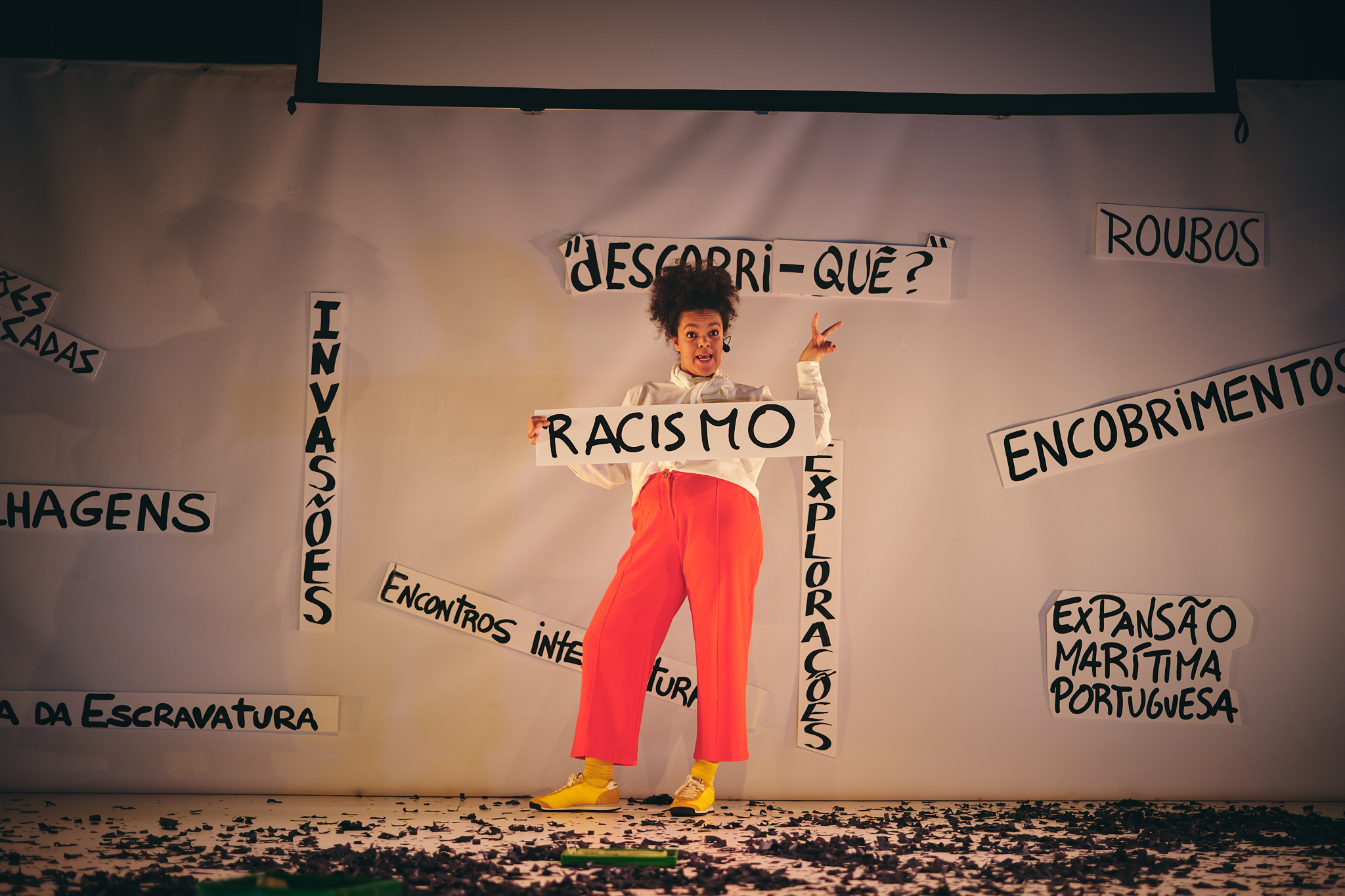 Uma mulher negra fala e gesticula de forma expressiva enquanto segura uma placa de papel branco com a palavra “RACISMO”. Atrás dela, uma tela branca exibe várias placas com palavras como “INVASÕES”, “ROUBOS” e “ENCOBRIMENTOS”.