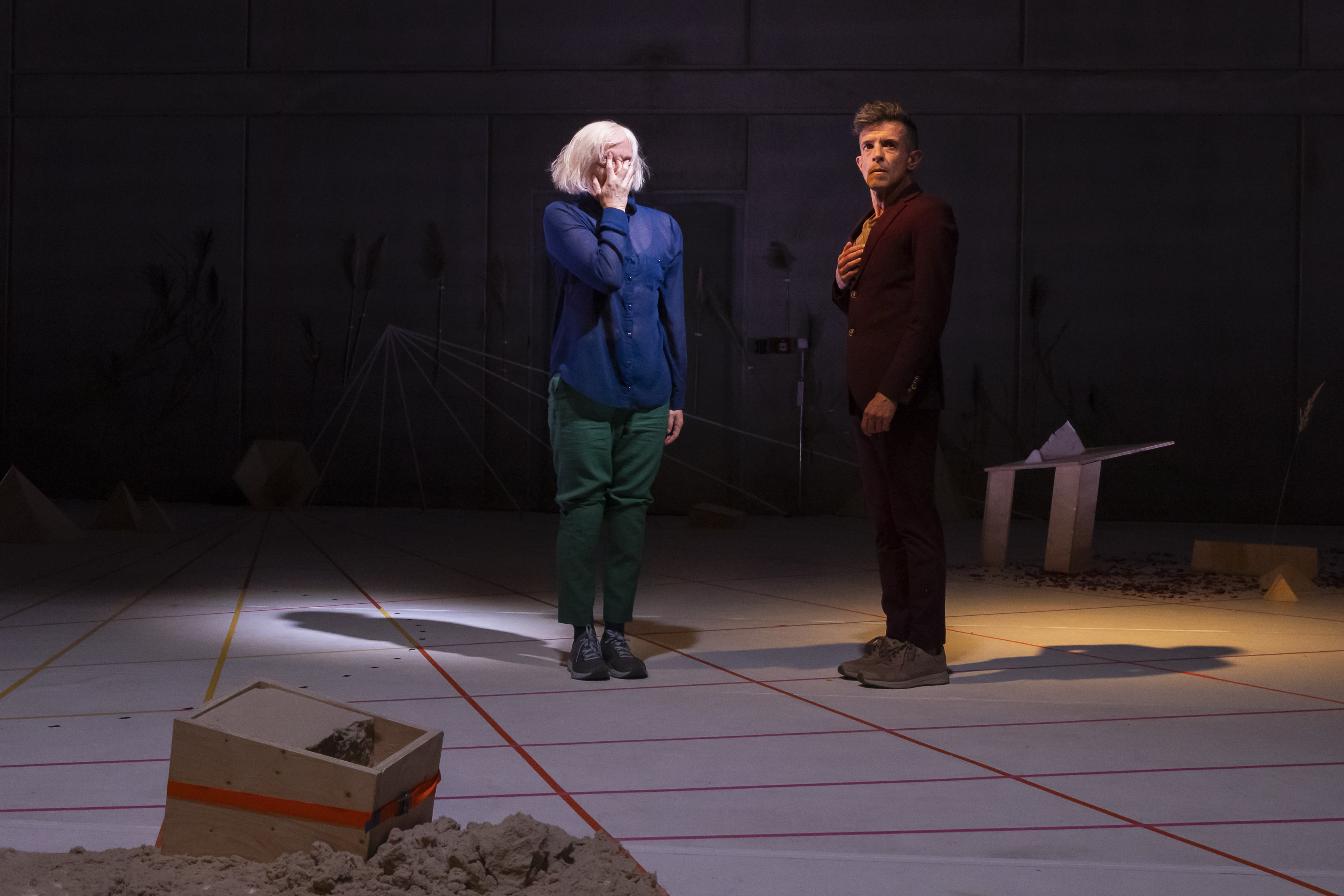 Color scene photo. Meg Stuart and Francisco Camacho are standing on a white floor with red lines forming squares on it. Meg covers her face. Francisco has one hand on his chest. 