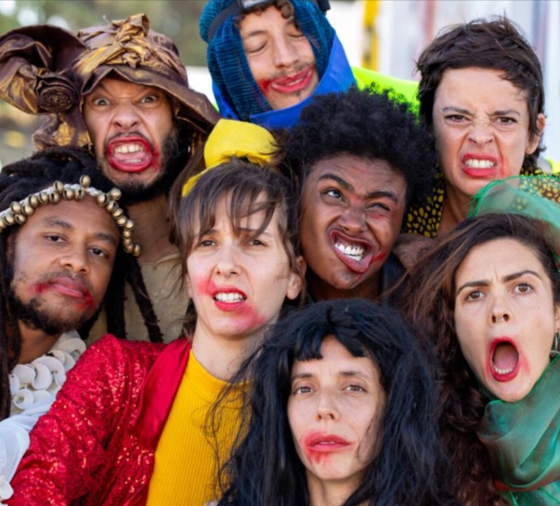 Fotografia a cores. Um grupo de intérpretes posa muito próximo, preenchendo o enquadramento com expressões exageradas e maquilhagens borradas. Os figurinos são variados e vibrantes, criando um conjunto visual caótico e expressivo.