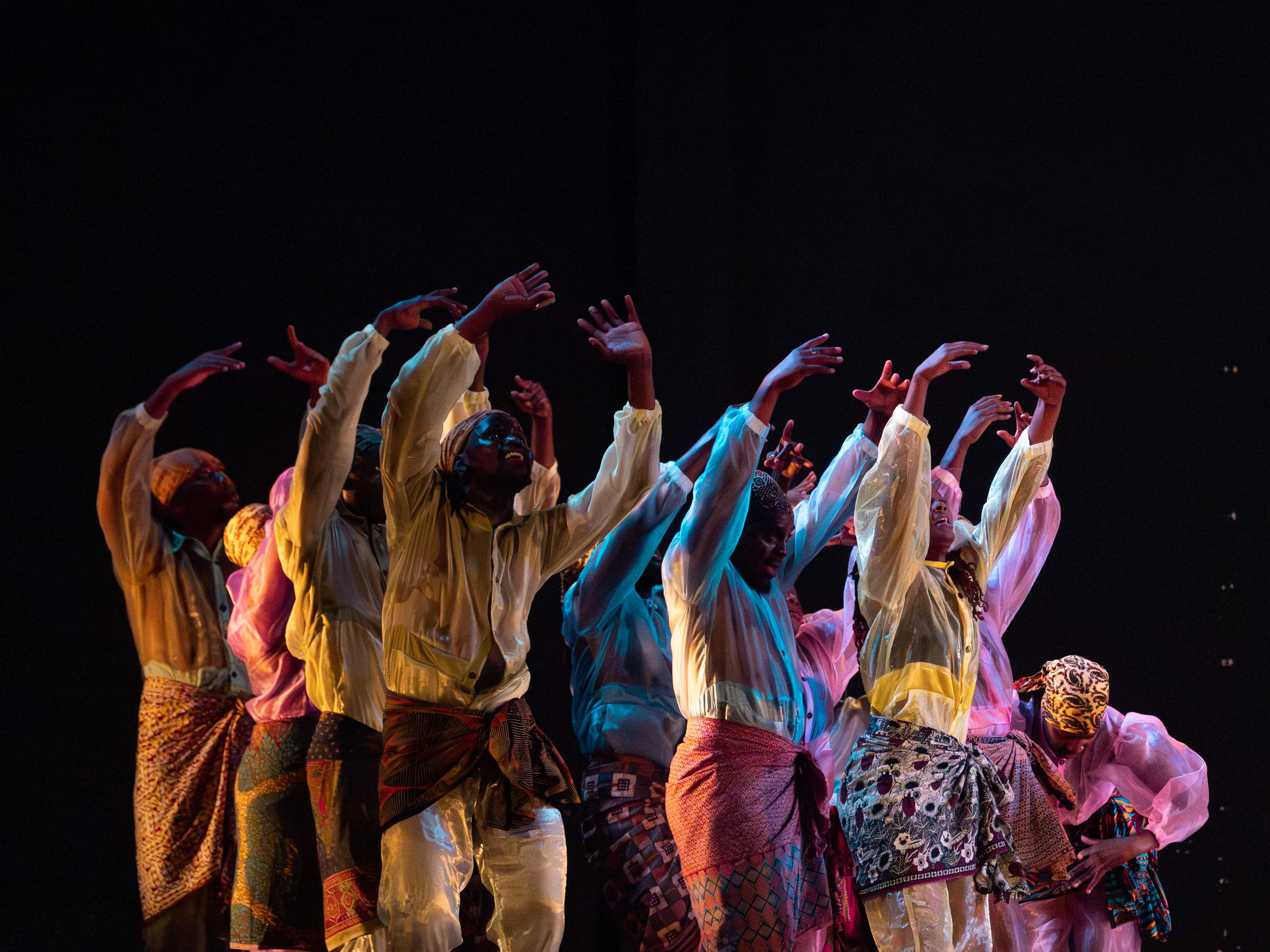A group of performers, wearing translucent, patterned costumes, raise their arms in a collective movement.