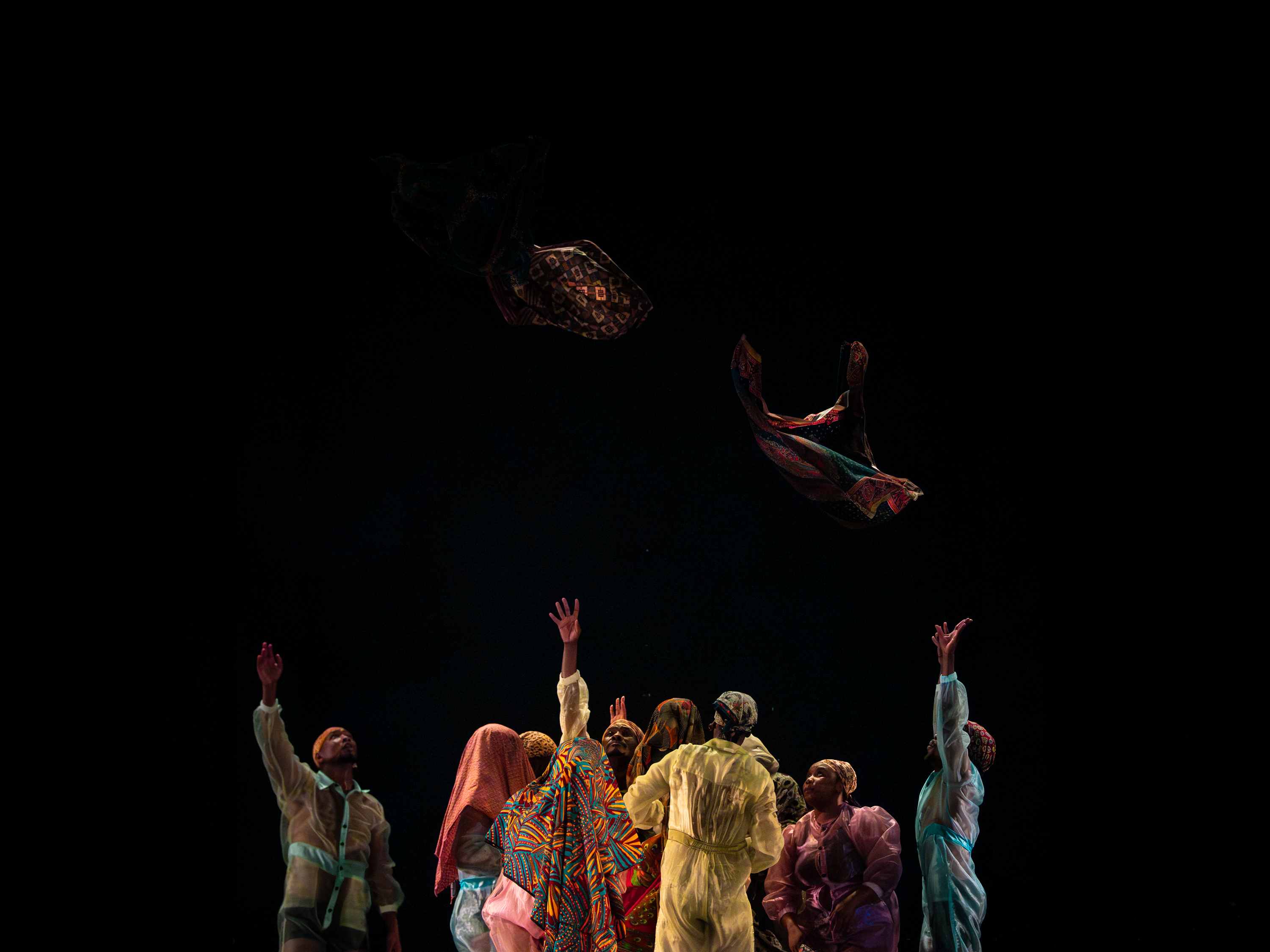 A group of performers throw patterned fabrics aloft during a scene, with their arms outstretched.