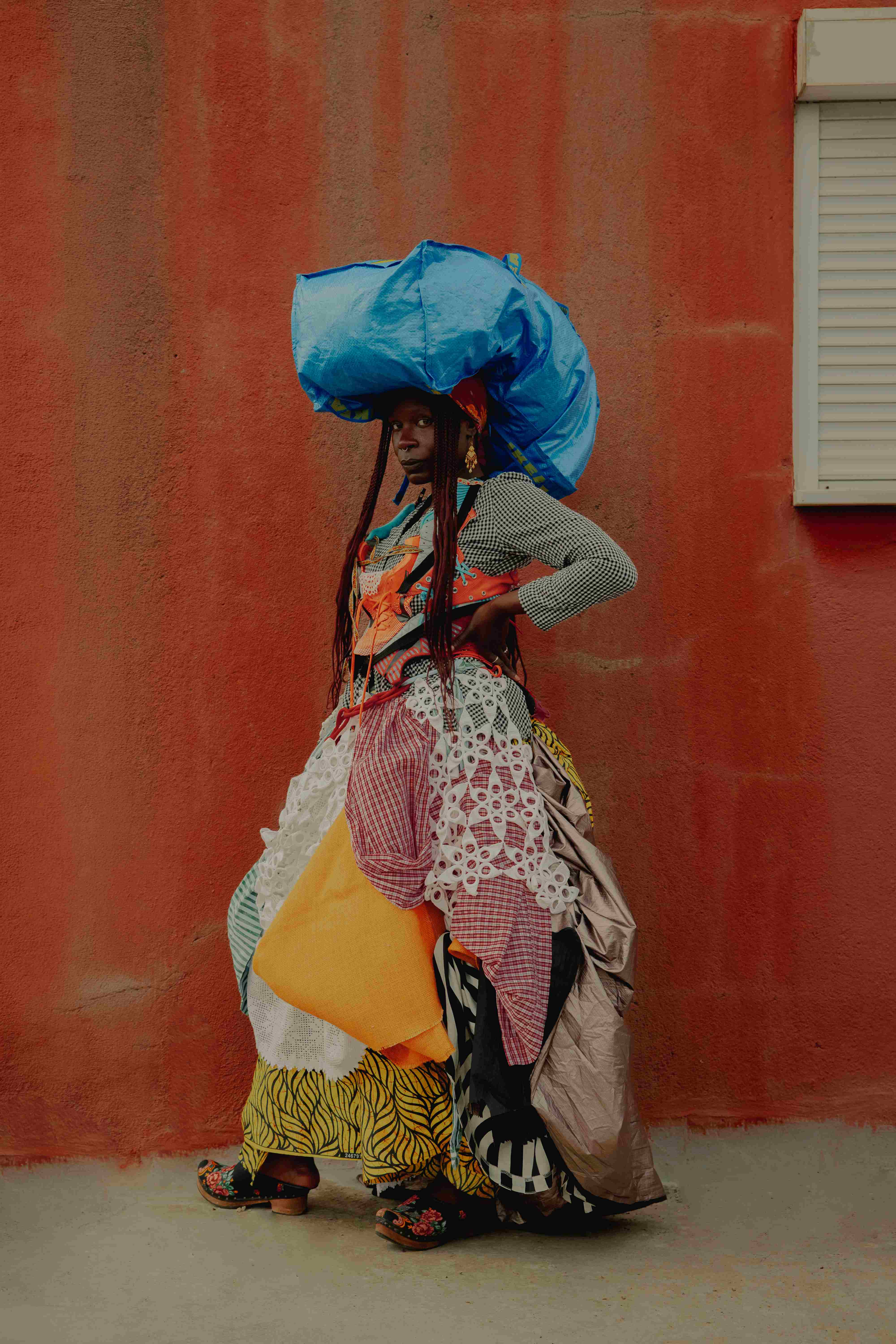 A woman poses in front of a reddish wall, wearing a garment made of colourful fabrics and balancing a large blue bundle on her head.