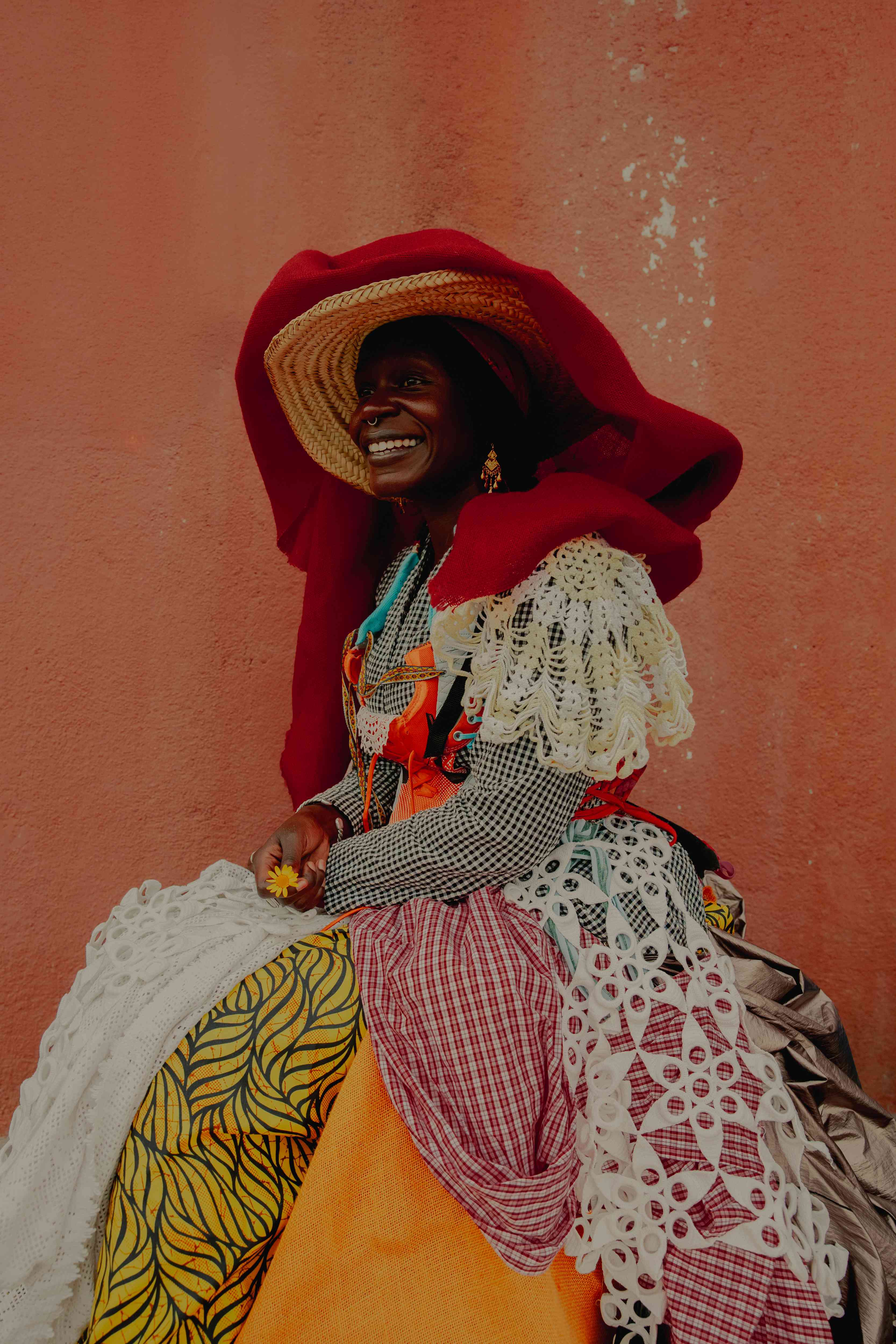 Standing in front of a reddish wall, a woman smiles, wearing a straw hat wrapped in red fabric and a dress made of colourful fabrics.