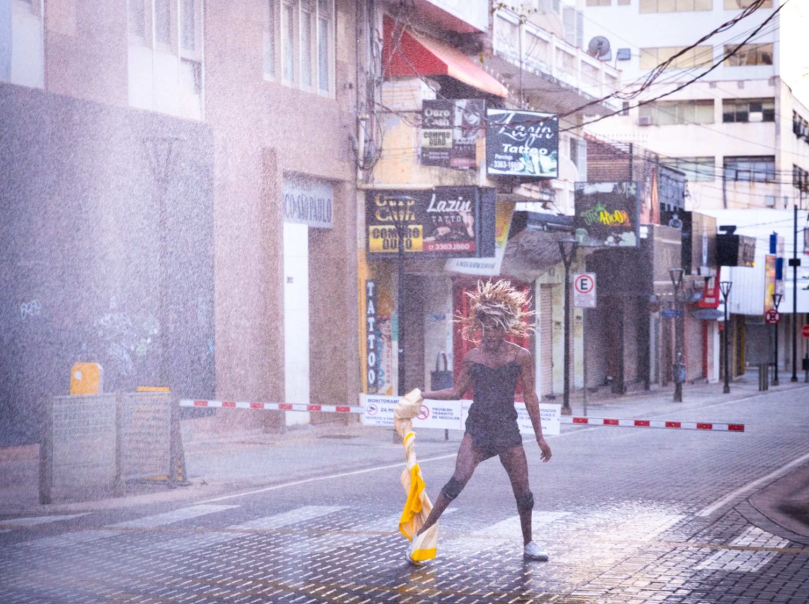 A person is standing in the middle of a wet street, holding a yellow and white piece of fabric, whilst a jet of water cuts across the scene.