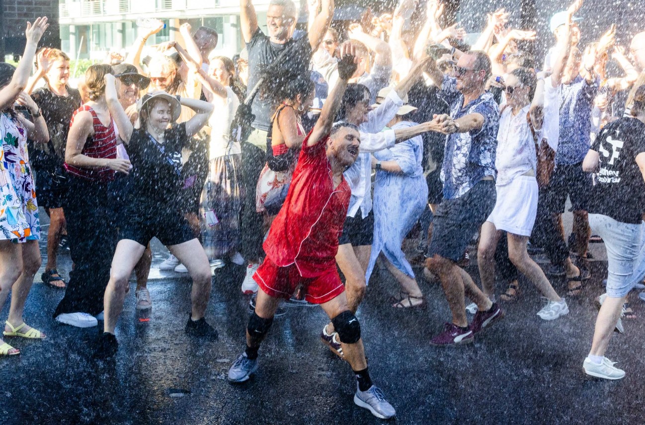 A group of people are dancing and raising their arms whilst being sprayed with jets of water on an open-air street.