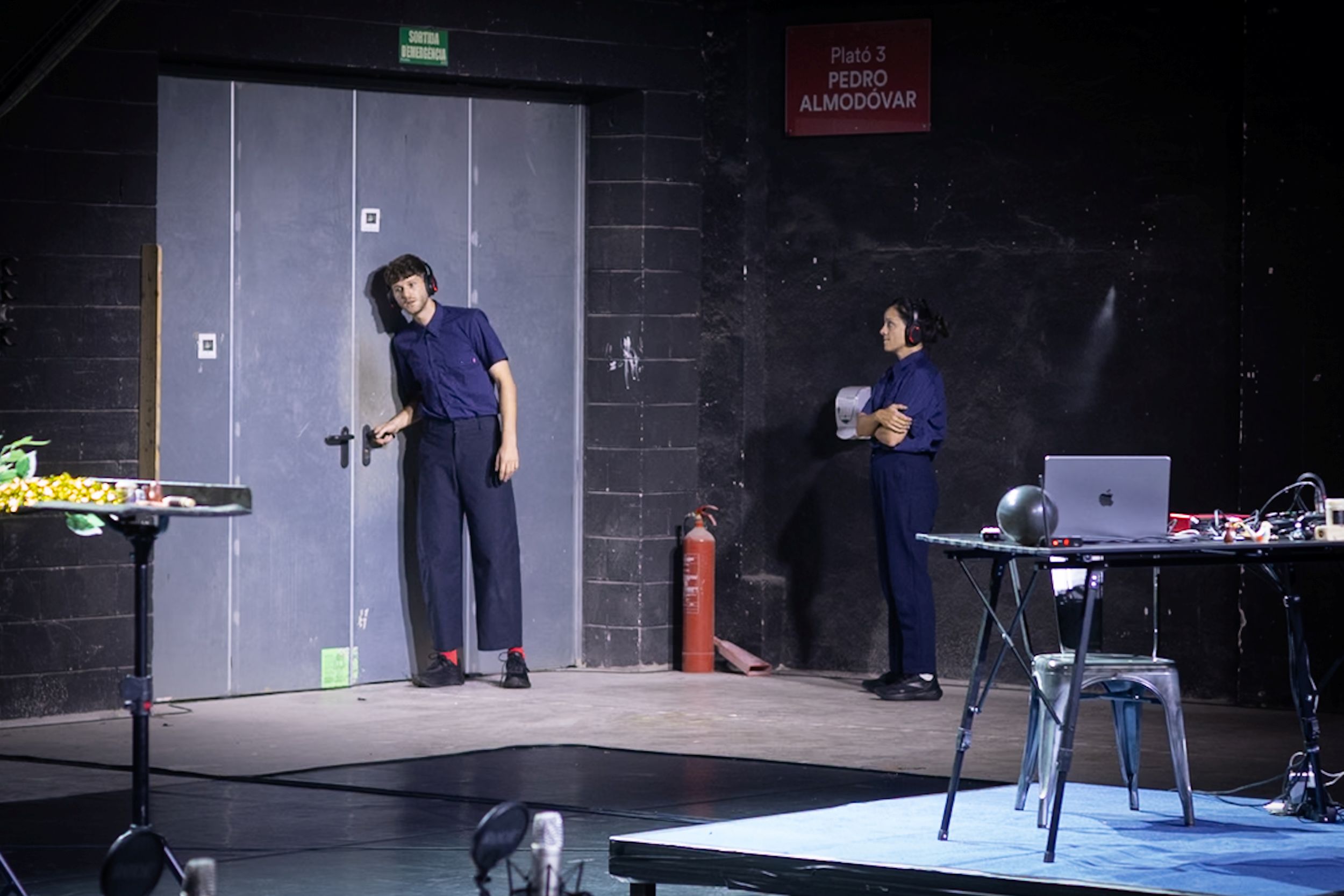 Two performers wearing headphones interact on stage, whilst one peeks through a door and the other watches from beside a table laden with technical equipment.