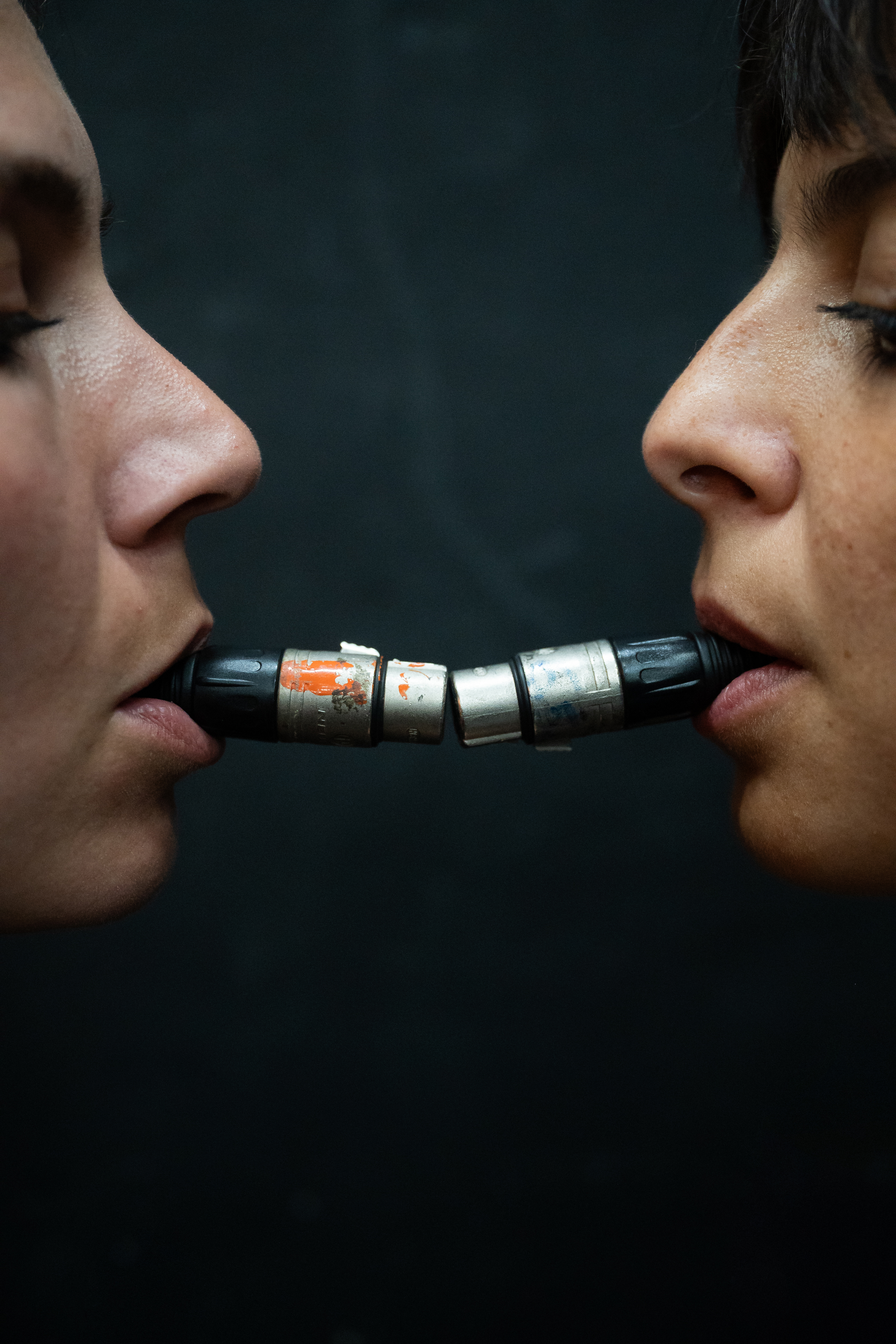 A close-up of two people facing each other, each with the end of a microphone lead in their mouth. The ends touch lightly.