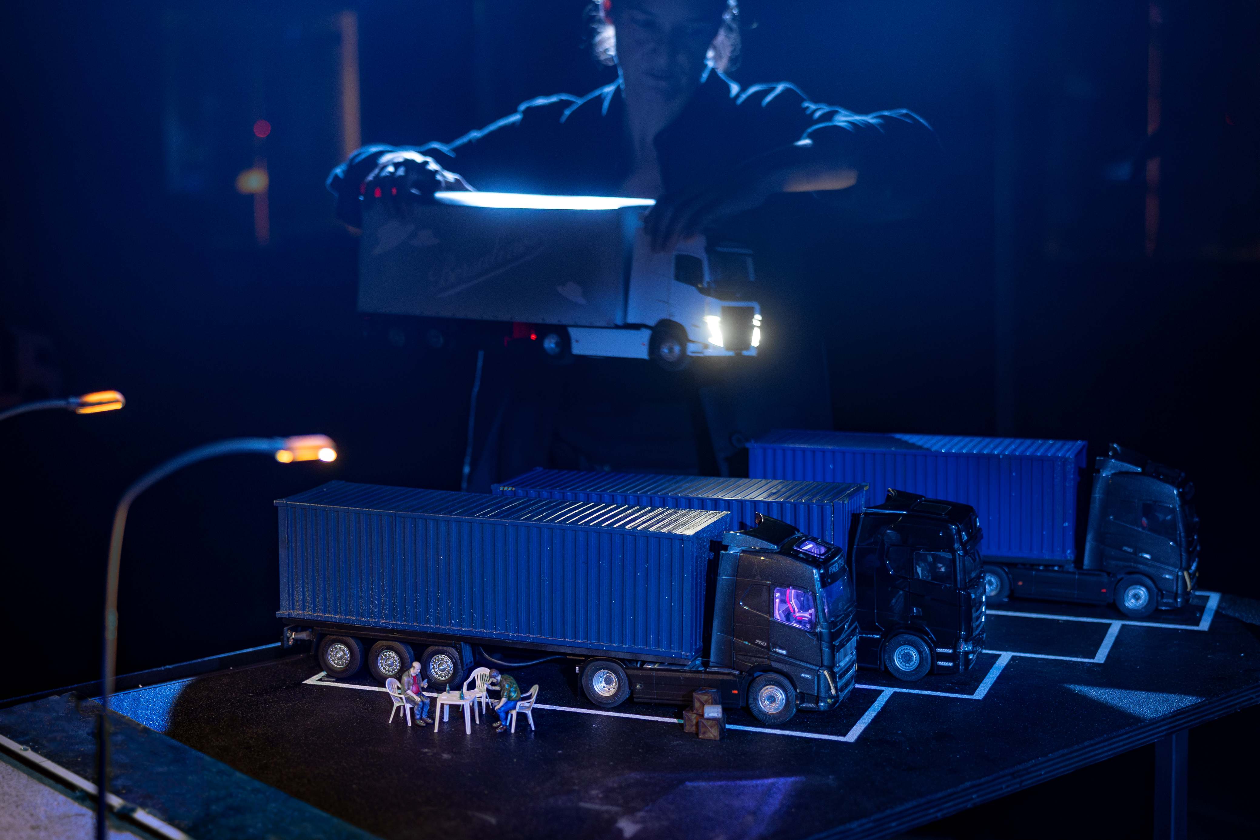 A performer moves a miniature lorry across a model featuring parked lorries.