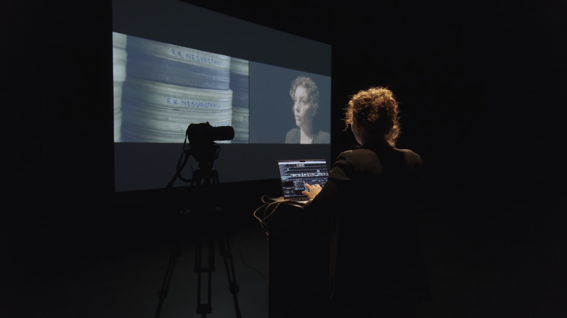 Color photograph. In a dark room lit only by a screen displaying a split projection — on one side, an image of film reels; on the other, Mila Turajli?’s face. Mila stands with her back to us, in front of a computer, with a camera pointed at her.