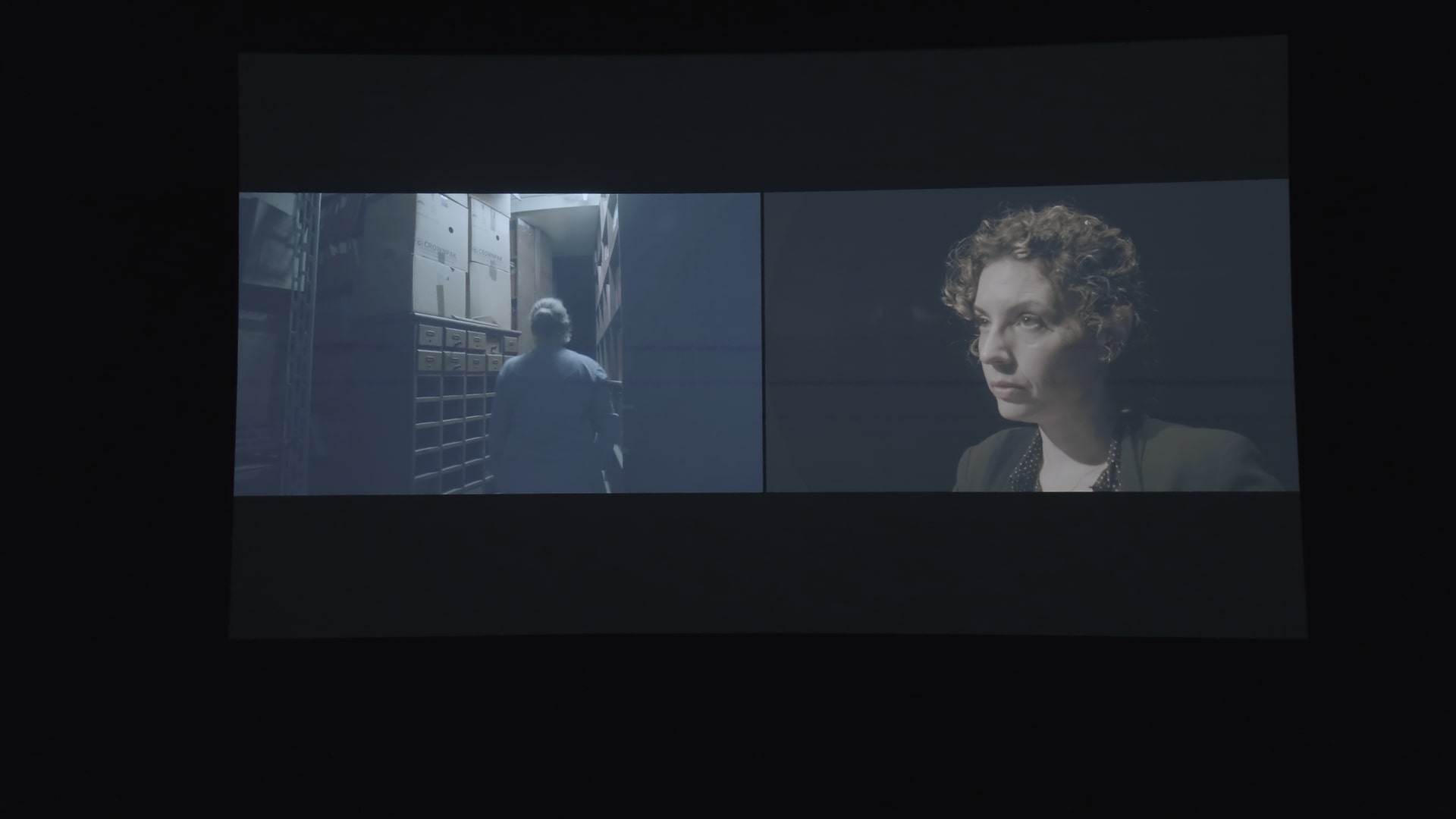 Color photograph of a screen in a dark room, showing a split display: one side features a person with their white hair tied back in an archive, and the other shows the face of Mila Turajli?.