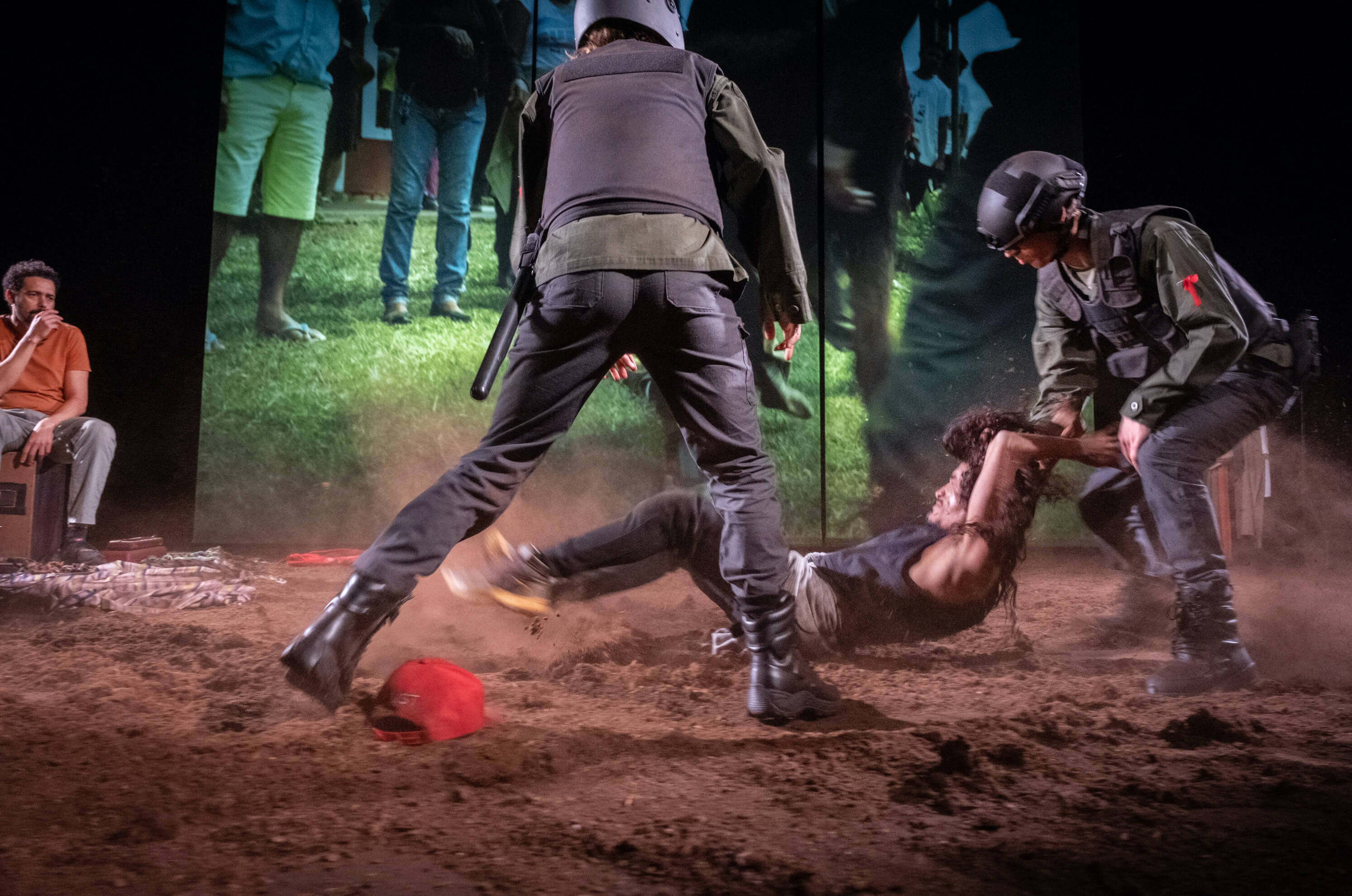 Fotografia de cena a cores. Dois intérpretes vestidos de polícias arrastam um terceiro intérprete pelo chão coberto de terra. 
