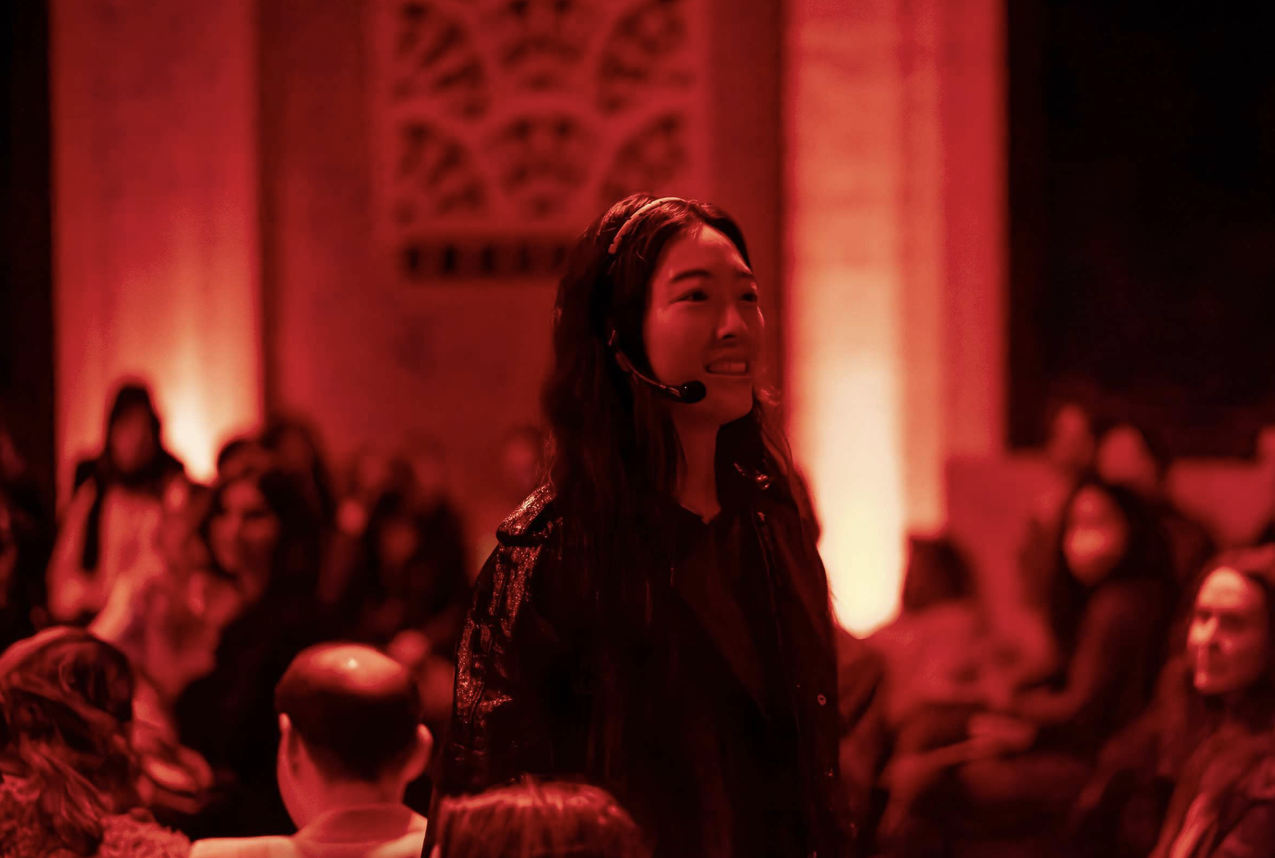 A person wearing a headset walks amongst the audience, bathed in red light, in an indoor space.