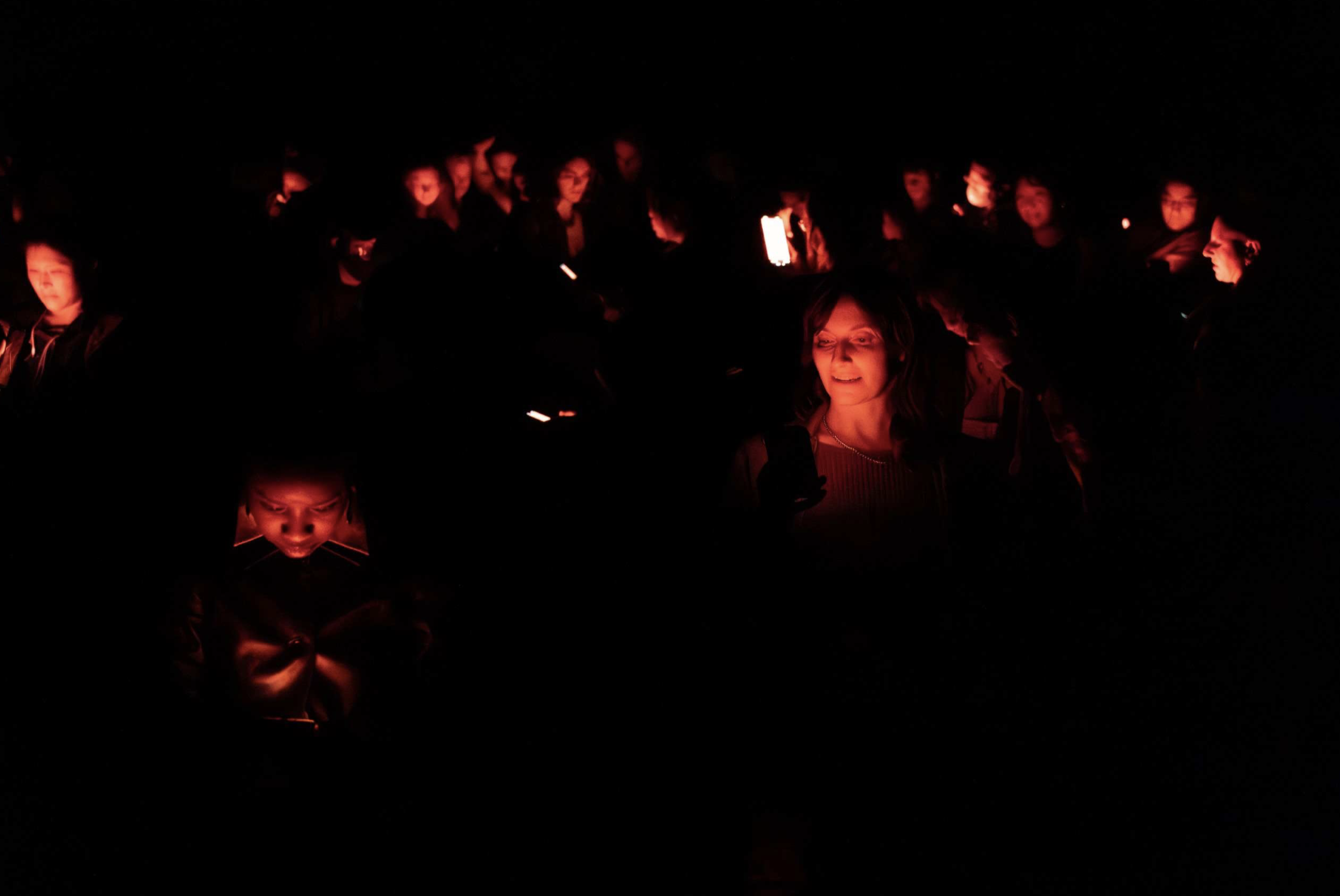 A group of people gather in a dark room, lit only by the light from the mobile phones they are holding.