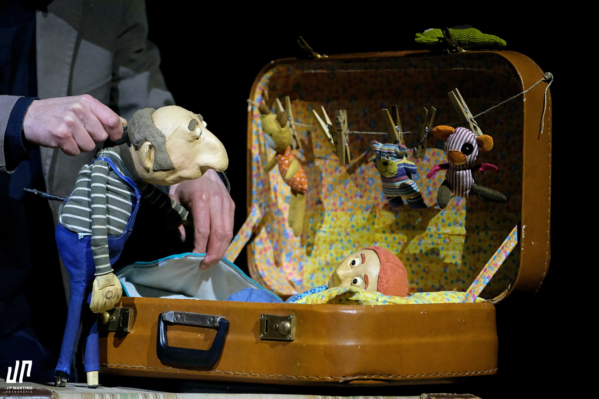 A puppet leans over an open suitcase in which another puppet is resting, surrounded by small hanging toys.