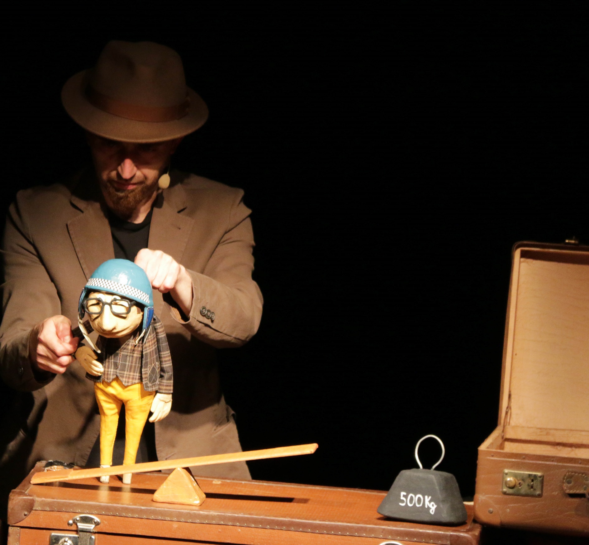 A puppeteer operates a puppet with a blue helmet balanced on a lever next to a 500 kg weight.