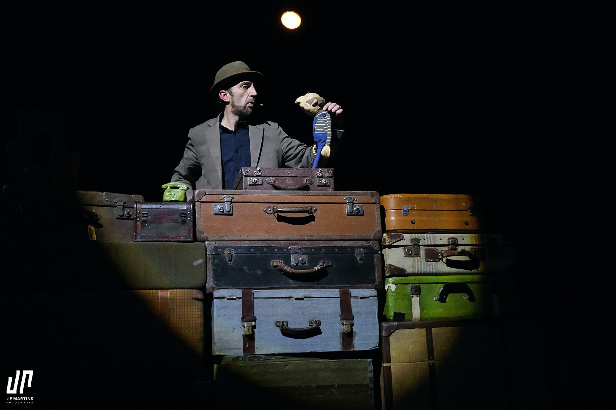 A performer interacts with a puppet on top of a pile of suitcases, illuminated by a spotlight, with the moon in the background.