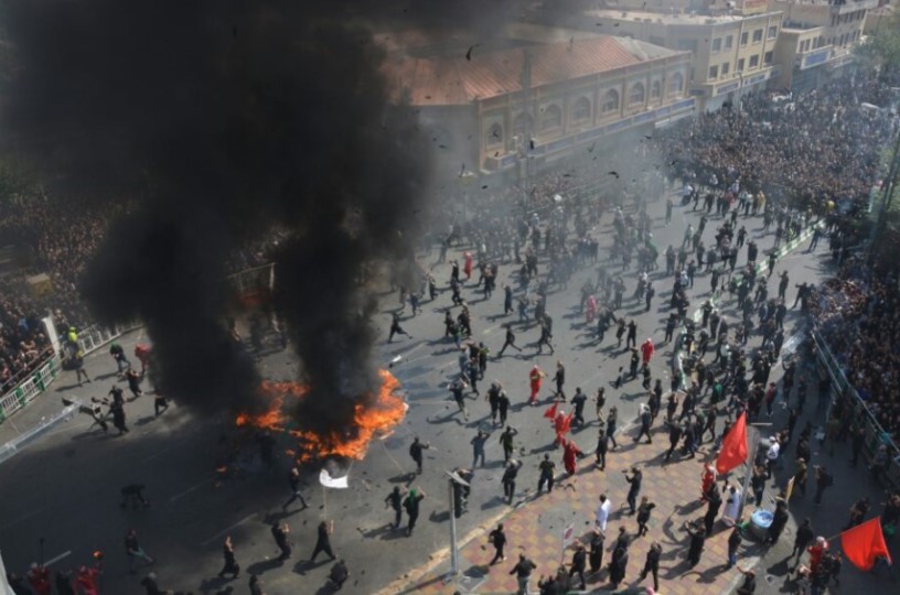 Fotografia a cores. Vista aérea de uma praça durante um protesto, com várias pessoas. No centro, objetos em chamas libertam fumo negro.