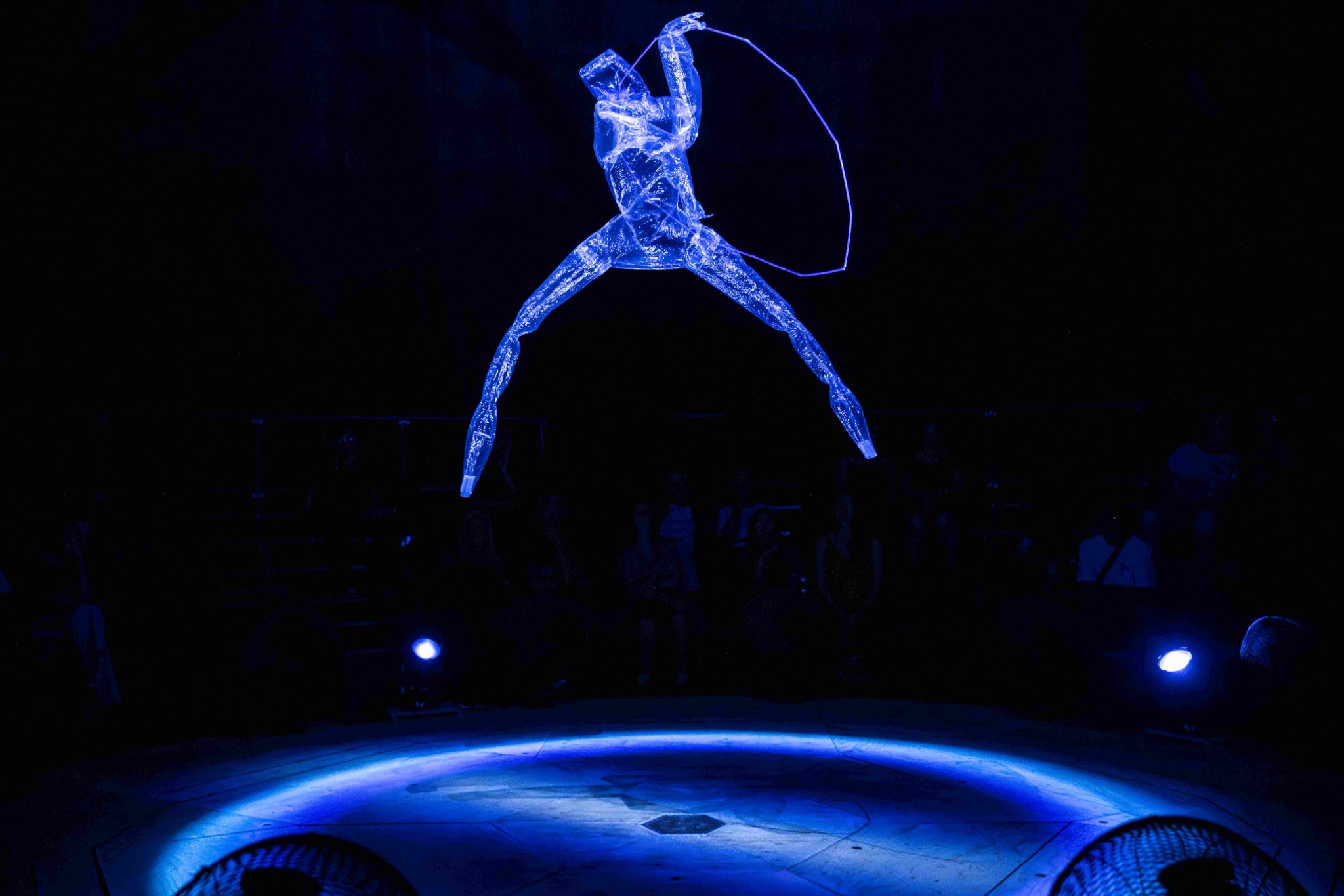 A translucent, anthropomorphic figure suspended in the air, with a hoop, illuminated by blue light.